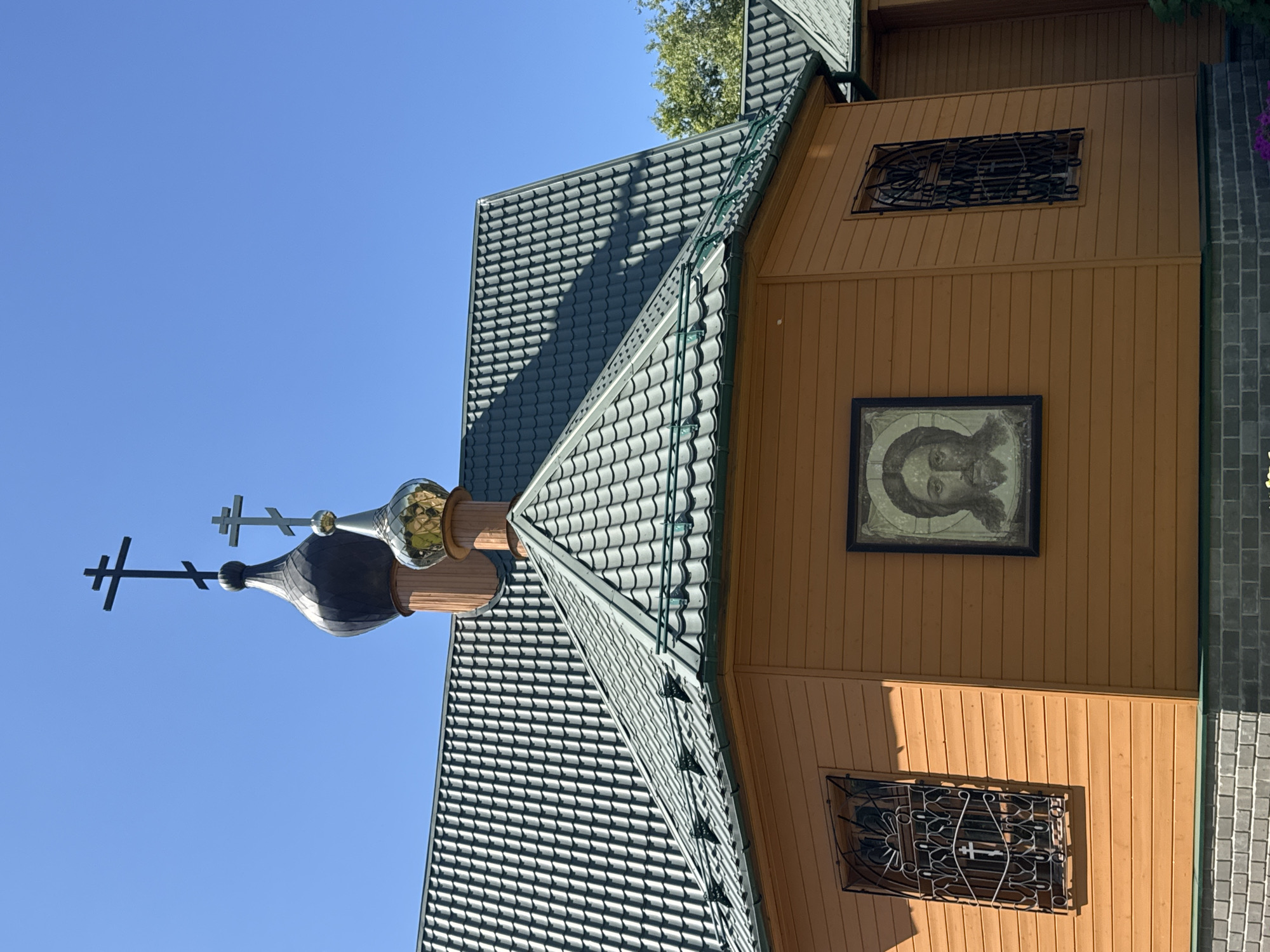 The image shows a view of a wooden church with a dark metal roof and a golden dome. Two crosses are visible on top of the church's domes. The church is decorated with an icon of Jesus Christ and features barred windows. The sky is clear and blue. The location is identified as Nizhny Novgorod, Russia.