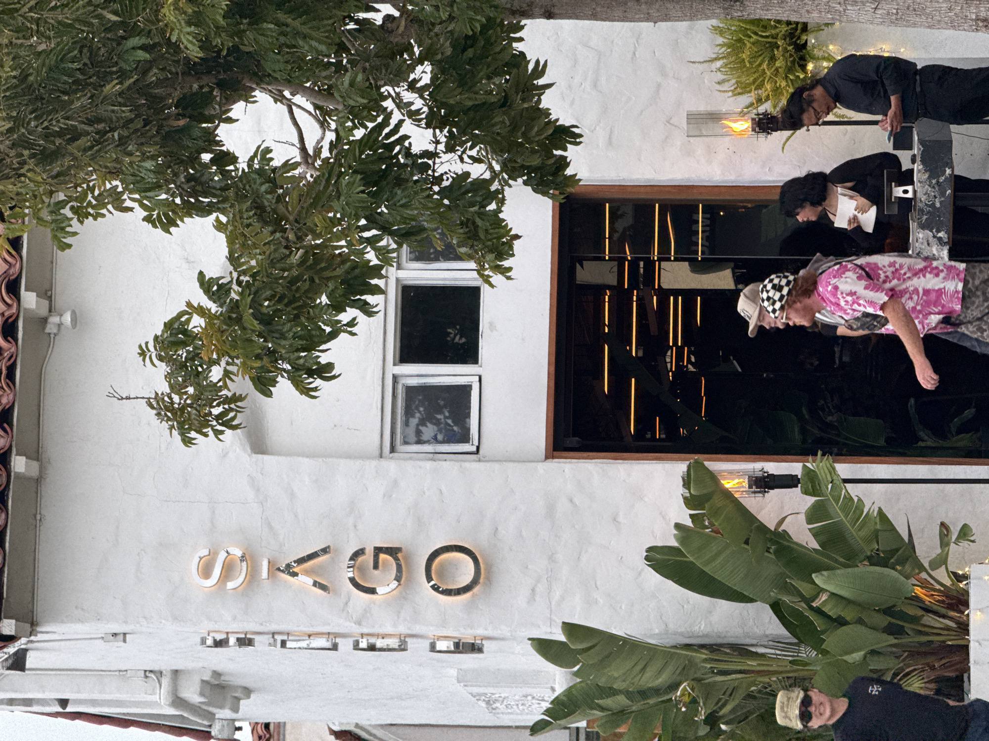 The image depicts the exterior of a white, textured building, identified as being in Encinitas, United States of America. A vertical sign on the left side of the facade spells "SAGO" with individual, illuminated, metallic-edged capital letters. Above the signage, a red-tiled roof is partially visible. Two small, dark, rectangular windows are located on the upper right portion of the building. A dark, recessed entrance, appearing to be glass, is situated on the lower right, revealing horizontal warm-toned interior lighting strips.
A large tree with green foliage stands on the right side, partially obscuring the building. On the left, a planter box contains large, broad-leafed green plants (possibly Strelitzia nicolai). Two black, vertical outdoor heating units with visible flames are positioned: one between the plants on the left and the building, and another to the right of the building's entrance.
Four individuals are present at the base of the building. From left to right:
1. A person wearing a camouflage cap, sunglasses, and a dark t-shirt with a white emblem, facing forward with a slight smile, partially visible.
2. A person with light-colored hair, wearing a black and white checkered cap, sunglasses, and a pink and white patterned short-sleeved shirt, walking towards the right.
3. A woman with dark hair, wearing a black top, standing behind a dark counter. She holds a white notepad and pen, appearing to look towards the person in the pink shirt.
4. A man with dark hair pulled back, wearing a black long-sleeved shirt and black pants, standing behind the counter and looking down at a small device held in his hands.
A small electronic device, possibly a point-of-sale system, is visible on the dark counter. A green, bushy plant in a white pot is positioned behind the man in black. The overall lighting suggests late afternoon or early evening.