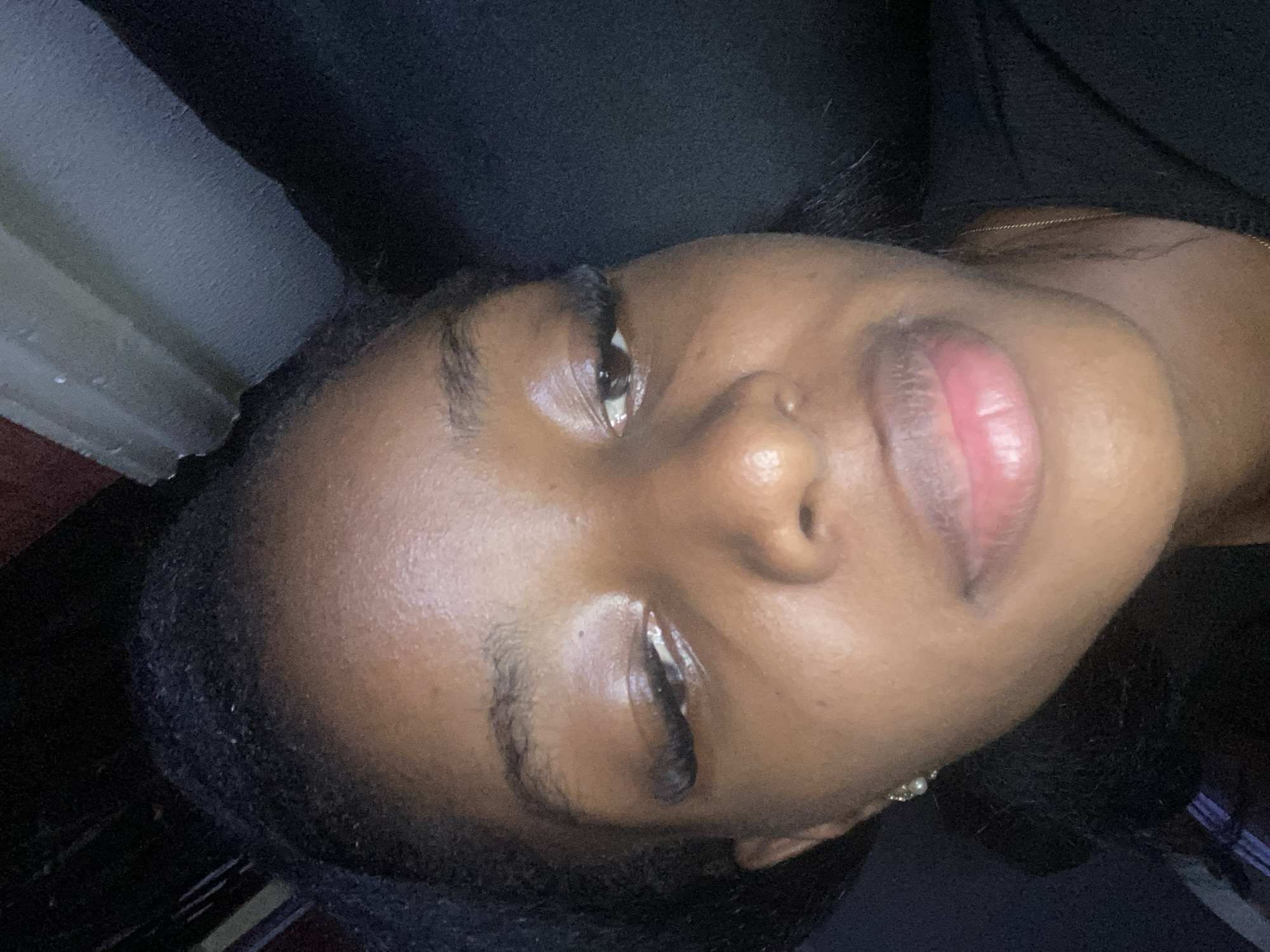 The image is a close-up, low-angle selfie of a young Black woman. She is looking upwards and slightly to her left, with her mouth slightly open. Her skin is a deep brown, and she has long, dark eyelashes. She is wearing a black top and a small, pearl earring in her left ear. The background is dark and out of focus, with hints of textured fabric or upholstery. The lighting is dim, with a warm tone. There is no discernible city or geographical information present in the image.