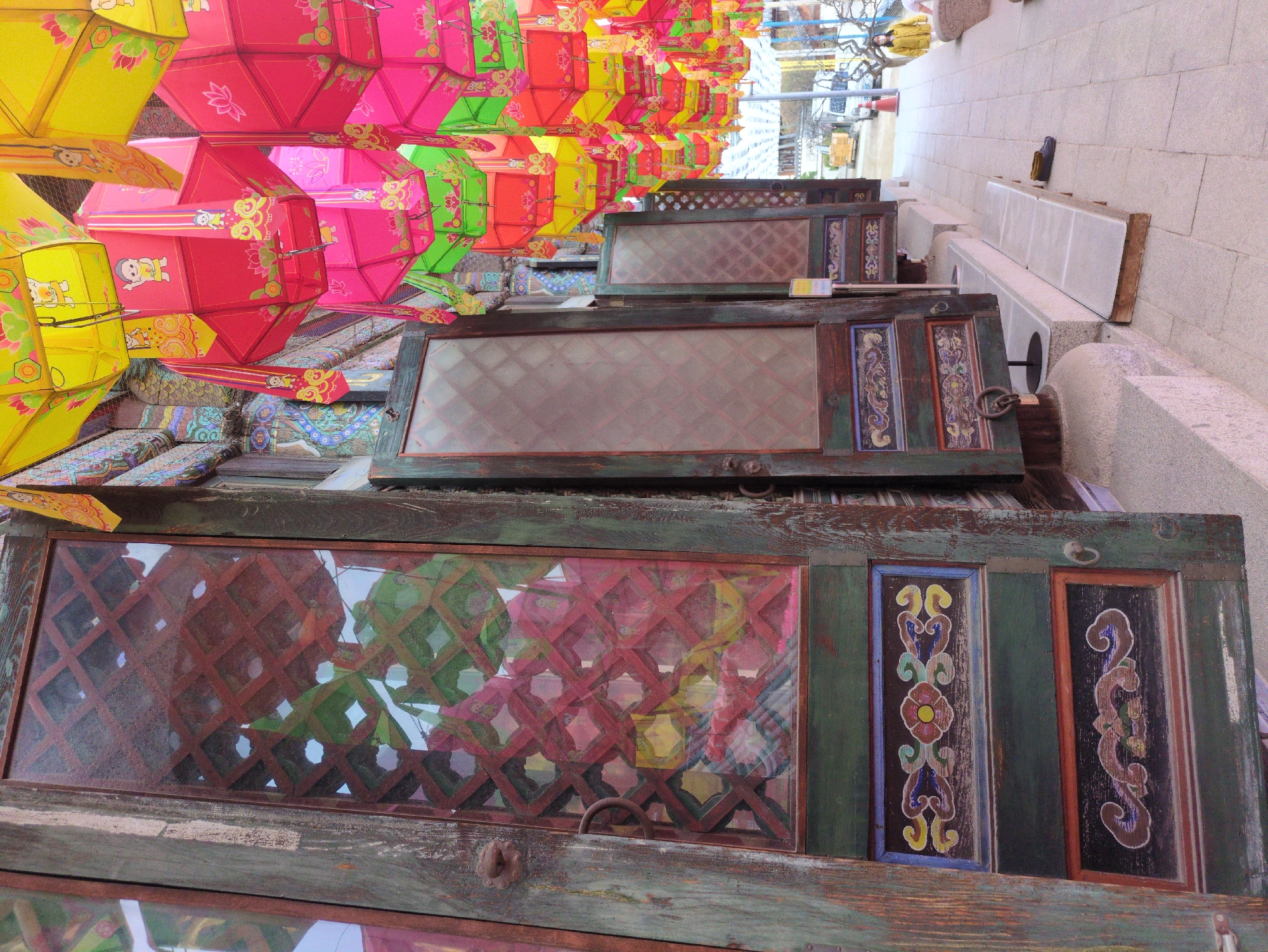 The image shows stacked wooden doors with glass panes propped against a light-colored stone wall in Seoul, South Korea. The doors are old, with weathered paint and decorative panels featuring floral and scroll patterns. Lanterns, colored in yellow, pink, red, and green, hang overhead, their reflections visible in the glass panes of the doors. The lanterns appear to be part of a street decoration or festival display. The background shows a street scene with buildings, traffic lights, and trees.