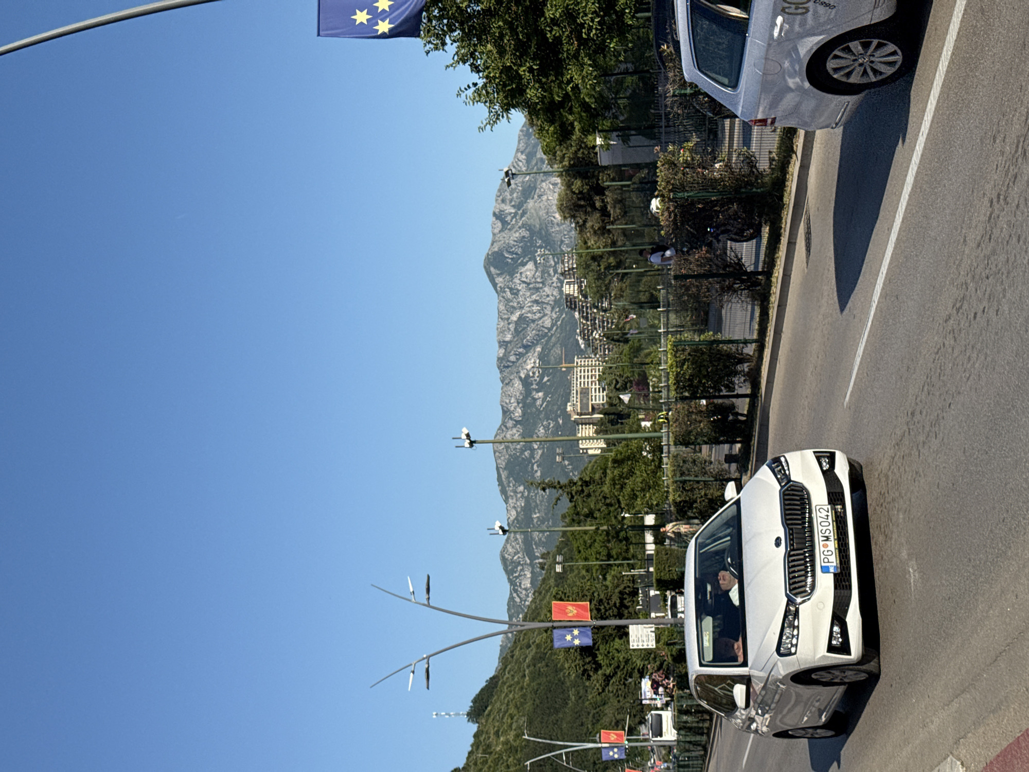This image captures a wide-angle daytime street scene in Budva, Montenegro, under a clear blue sky.
In the immediate foreground on the left, a white Skoda Fabia hatchback (likely a 2021 or newer model) is in motion, facing the viewer. Its Montenegrin license plate reads "PG MS042". Two individuals are visible inside the vehicle; the driver wears a light-colored hat or is bald. On the right, the rear portion of a white station wagon (possibly a Volkswagen Passat) is visible, showing its right rear wheel and a side door with partial text "GOL" and numbers "06830".
The road is asphalt, marked by a solid white dividing line. A red curb is present along the left edge of the road. Both sides of the road are lined with green hedges and trees, creating a dense barrier between the road and adjacent areas. Behind the hedges on the right, a dark motorcycle or scooter is partially visible, along with several pedestrians. One person in a light-colored top is clearly visible behind the hedge.
The mid-ground features a series of distinctive modern streetlights with multiple arms. Flagpoles along the roadside display alternating flags of the European Union (blue with twelve yellow stars) and the national flag of Montenegro (red with a golden double-headed eagle). Further back on the left, a white bus is partially obscured by foliage.
The background is dominated by a range of large, rocky mountains, partially covered with trees and scrub, under a cloudless blue sky. Scattered multi-story buildings, some with terraced architecture, are visible on the lower slopes of these mountains. A cellular or radio tower is discernible on a distant hillside to the far left. The scene is brightly lit by direct sunlight, casting shadows.