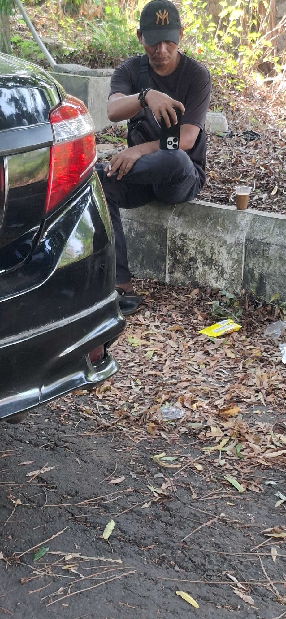 The image depicts a person sitting on a concrete ledge next to a black car. The person is wearing a dark t-shirt, dark pants, and a black baseball cap with a white logo. They are holding a smartphone in their right hand and a cigarette in their left hand. The car, a black sedan, is parked on the left side of the frame, with its rear taillight visible. The ground is covered with dry leaves and dirt. In the background, there are trees and foliage. A clear plastic cup containing a dark liquid is placed on the ledge to the right of the person. The scene appears to be an outdoor setting, possibly a street or parking area in Cilaku, Indonesia, characterized by its natural environment with fallen leaves and vegetation.
