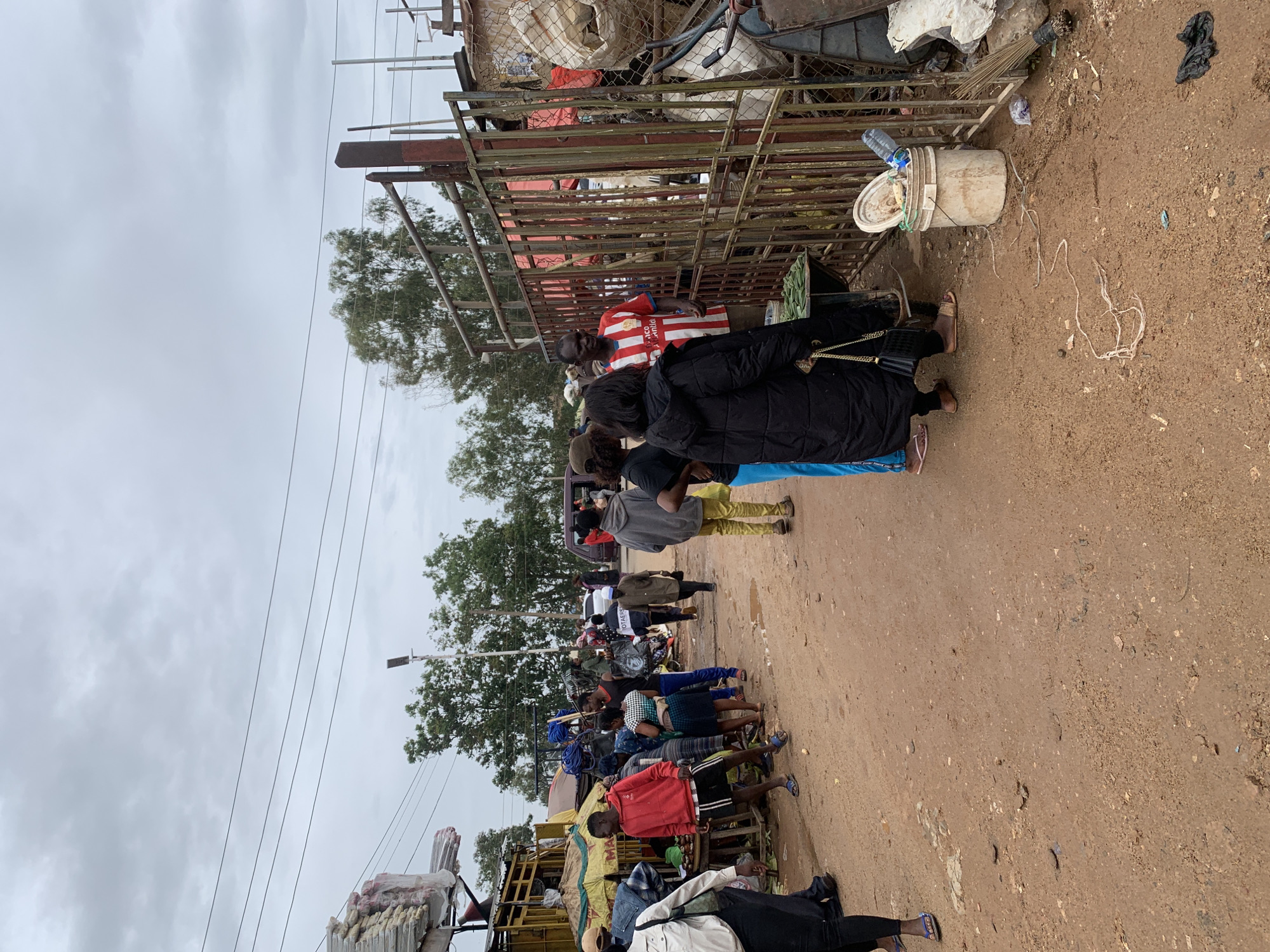 The image depicts a dirt road scene in Gura Top, Nigeria. A line of people stands along one side of the road. They are mostly positioned near makeshift stalls or carts piled high with goods. The individuals are dressed in casual clothing. A metal fence or structure runs along the edge of the scene. Overcast sky suggests a cloudy day.