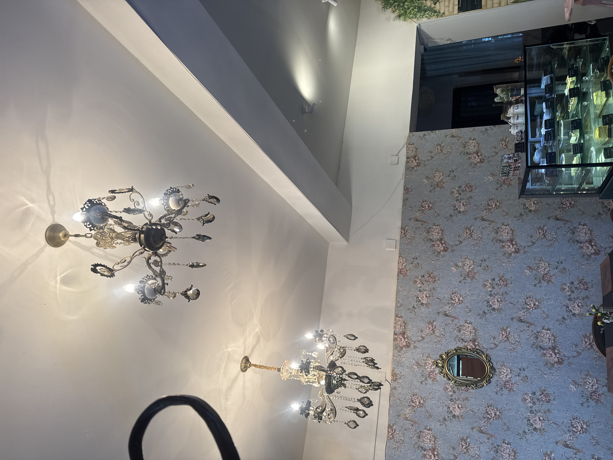 A low-angle interior shot reveals a room with two chandeliers illuminating its space. The wall is adorned with a floral pattern. A mirror is mounted on the wall. A display case, filled with items, occupies a corner of the room. A segment of what appears to be a black object is in the foreground.