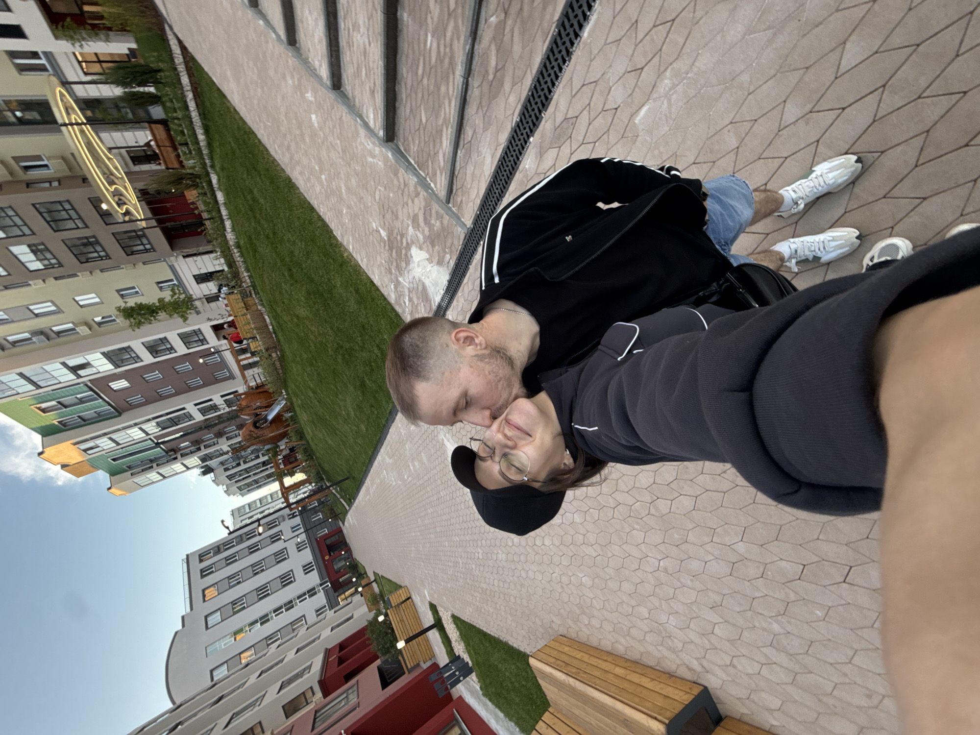 This image captures a tender moment between a couple in a modern residential area of Pervomaysky, Russia, during what appears to be late afternoon or early evening. The photo is taken from a slightly upward, selfie-like angle.
In the foreground, a male with short, light brown hair and a beard, dressed in a black track jacket with white stripes (displaying a small white 'M' logo), denim shorts, and white sneakers, is seen kissing the forehead or cheek of a female. The female, wearing glasses, a black baseball cap, a dark grey/black hoodie, and white sneakers, has her eyes closed and a contented smile, indicating affection and happiness.
They are standing on a light-colored pedestrian path made of hexagonal tiles, bordered by well-kept green grass. The setting is an urban or suburban environment characterized by contemporary residential buildings of various heights and colors (white, grey, brown, green, and red accents) in the background. A clear, pale blue sky is visible above the buildings.
Notable details include decorative street lights, one of which features a prominent, abstract, glowing light sculpture. Wooden benches are placed along the pathway, and glimpses of a playground structure can be seen further in the distance. The soft lighting and the intimate pose of the couple convey a sense of warmth and quiet enjoyment in this pleasant, well-maintained public space.