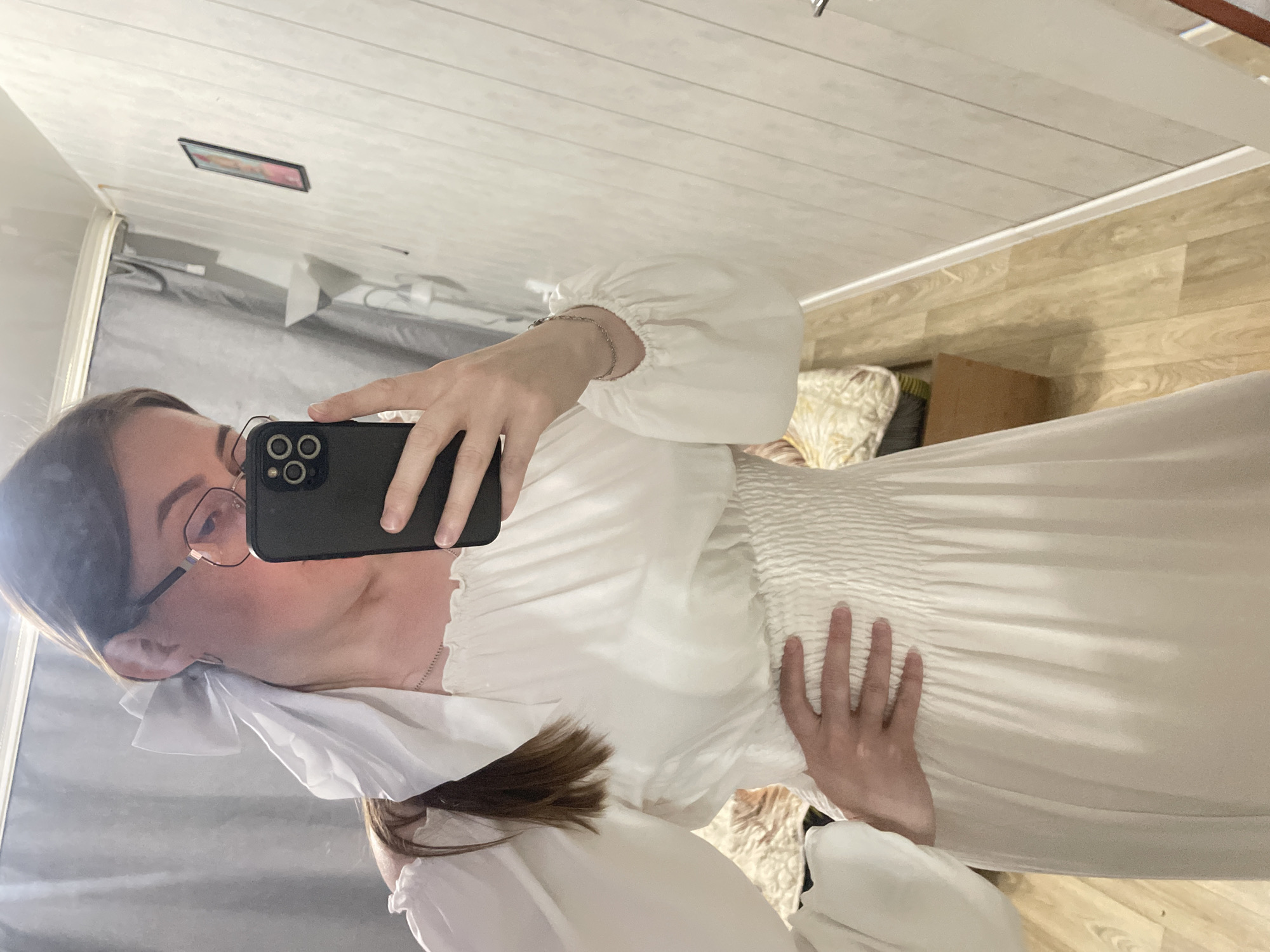 In the city of Sykrovo, Russia, a young woman with brown hair and glasses is taking a mirror selfie in what appears to be a room. She is wearing a white dress with a smocked bodice and voluminous puff sleeves. A white bow is tied in her hair. Her left hand is resting on her hip, and she is holding a black smartphone to take the picture. The room has wood-paneled walls and a wooden floor. A framed picture hangs on the wall, and a lamp is visible in the background. A bed with a patterned cover is also partially visible.