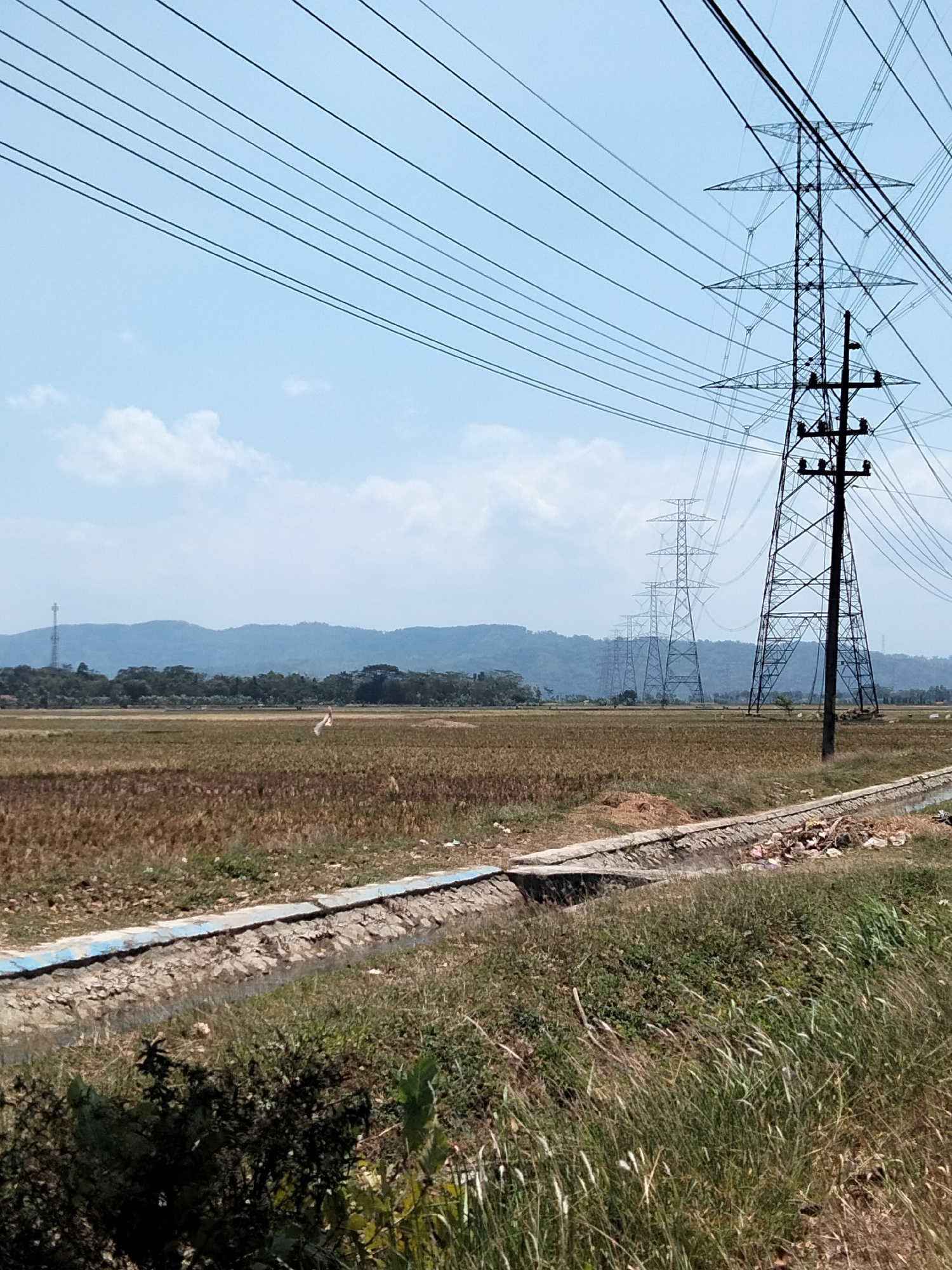 The image depicts a rural landscape in Karangkemiri, Indonesia, characterized by a clear blue sky with scattered clouds. Dominating the scene are several tall, metal electricity pylons carrying power lines that stretch across the frame. In the mid-ground, a dry, harvested rice field is visible, with patches of brown stubble. A thin, concrete-lined ditch runs along the foreground, partially filled with debris and water. Beyond the field, a line of trees and rolling hills form the horizon. A single, white object, possibly a piece of cloth or a kite, is seen fluttering in the distance within the field. The overall impression is one of open space and agricultural land under a bright sky, with a significant industrial infrastructure element.