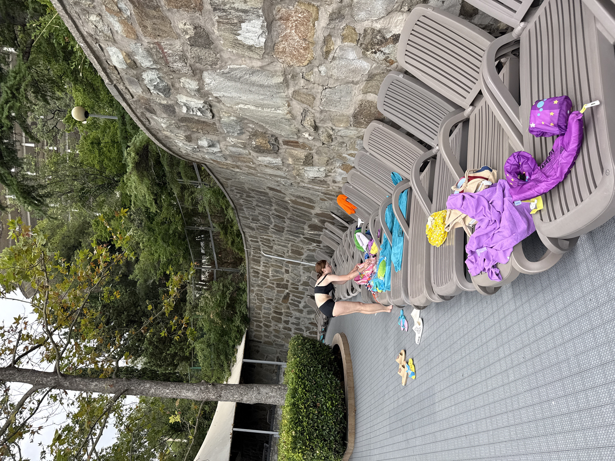 In the city of Massandrovsky Village Council, Ukraine, a woman in a black swimsuit bends over lounge chairs arranged on a gray tiled deck. She is sorting through a pile of colorful beach towels and clothing. Several pairs of flip-flops are scattered on the deck. Behind the lounge chairs, a textured stone wall curves along the property. Lush green trees and foliage line the top of the wall, and a vintage light fixture is visible among the branches. A large, established tree stands to the left of the scene, with its trunk partially obscured by a dense green hedge.