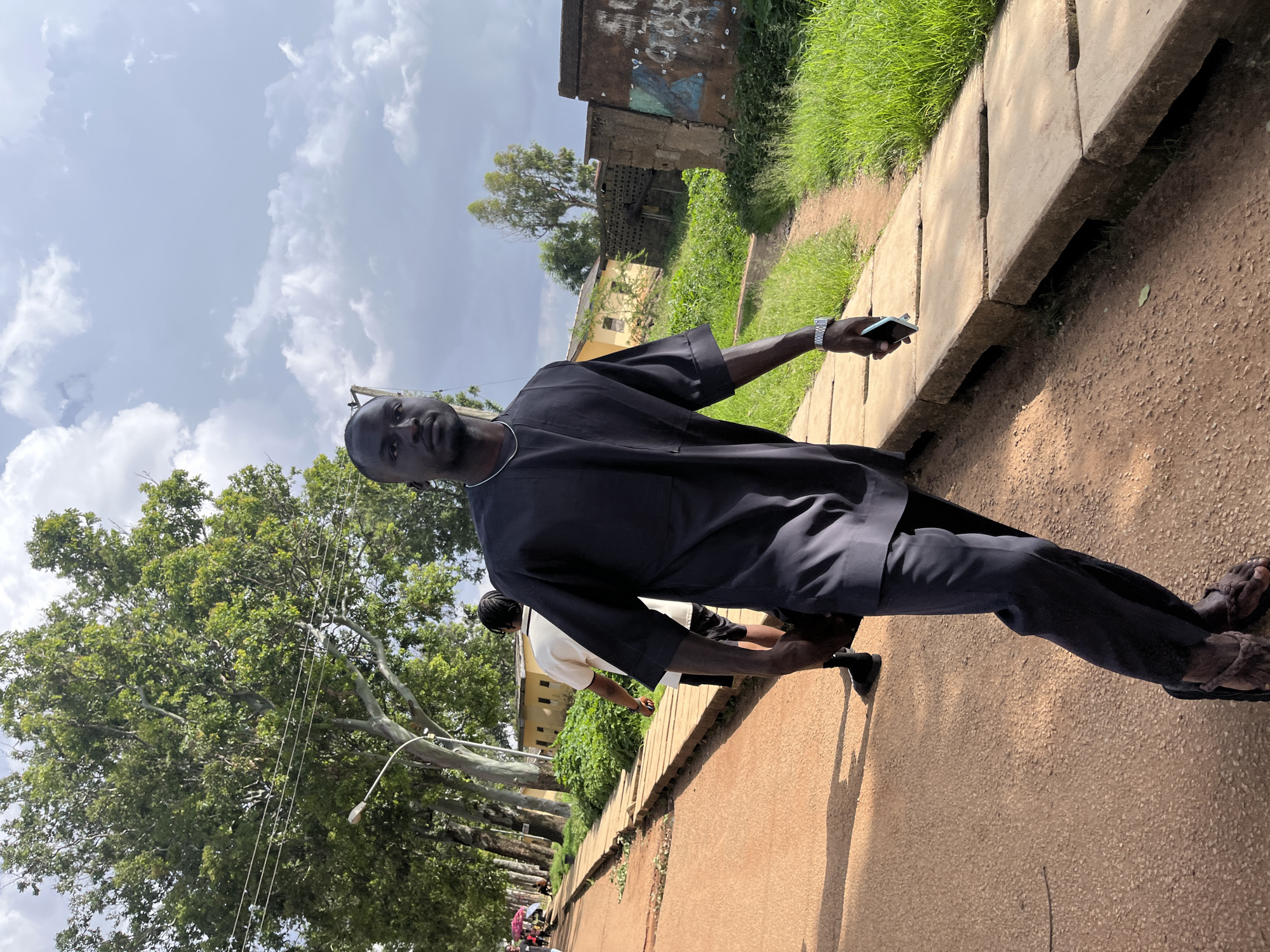 A Black man wearing a dark grey traditional outfit walks towards the camera on a dirt path lined with concrete slabs. He is on the right side of the frame, with his body angled slightly away. He has short dark hair and is looking forward. He is holding a light blue smartphone in his right hand. Behind him, to the left, another person wearing a white shirt and dark shorts walks away from the camera. In the background, there are buildings, lush green trees, and a cloudy sky. The scene appears to be in a semi-urban or rural area. The overall impression is of a casual outdoor setting.