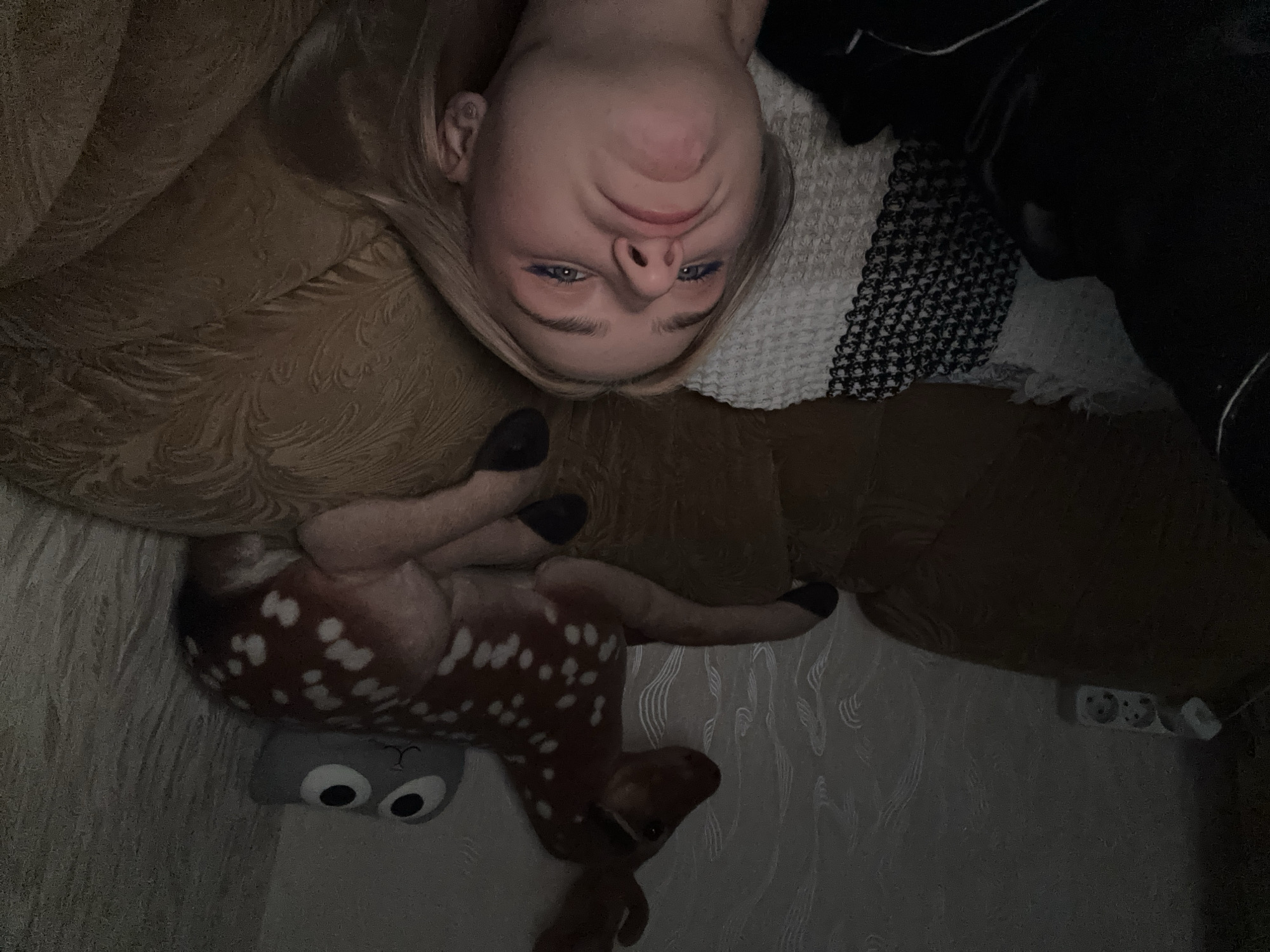 A young blonde woman with blue eyeliner is lying down on a couch, facing the camera with a pouty expression. She is wearing dark clothing and has earrings in her ears. Next to her, a stuffed deer with brown fur and white spots is lying on the couch, along with a grey stuffed animal with large eyes. In the background, an electrical outlet is visible on the wall, which is covered in patterned wallpaper. The scene is dimly lit, suggesting it is nighttime or indoors with minimal lighting. The overall environment appears to be a living room or bedroom.
