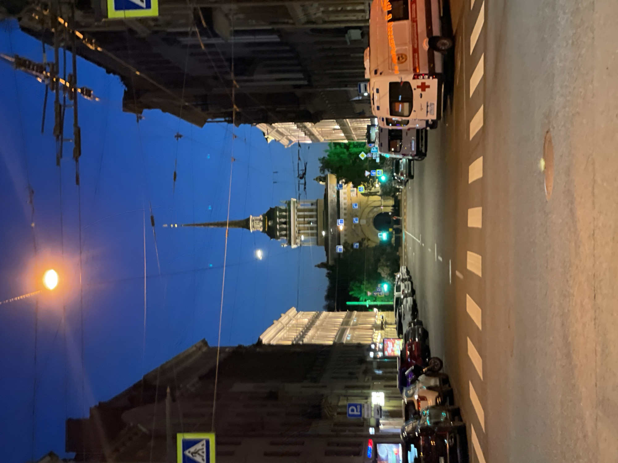 The image captures a twilight or nighttime scene on a wide city street in Saint Petersburg, Russia. The perspective looks down a long avenue towards a prominent architectural landmark.
In the distance, the iconic Admiralty Building with its distinctive tall, golden spire and arched gateway stands as the central focus, illuminated against the dark blue sky. Buildings with classical architecture, some featuring columns and illuminated facades, line both sides of the street.
On the right side of the road, a white ambulance with a red cross symbol and Cyrillic text reading "МЕДИЦИНСКАЯ СЛУЖБА" (Medical Service) is parked. Further down, other vehicles are dimly visible. The asphalt road features white zebra crossing stripes.
On the left side of the street, numerous cars and motorbikes are parked along the curb. A multi-story building with illuminated upper floors showcases its architectural details. Overhead, a complex network of power lines, likely for electric transport, is visible against the sky.
Artificial streetlights cast bright glows, with one particularly luminous circular light source in the upper left, while the illuminated buildings provide additional light. Visible street signs include a blue square sign with a white 'P' symbol for parking and a triangular sign with a blue background and a white pedestrian symbol, indicating a pedestrian crossing. The overall atmosphere suggests a bustling urban environment transitioning from dusk to night.