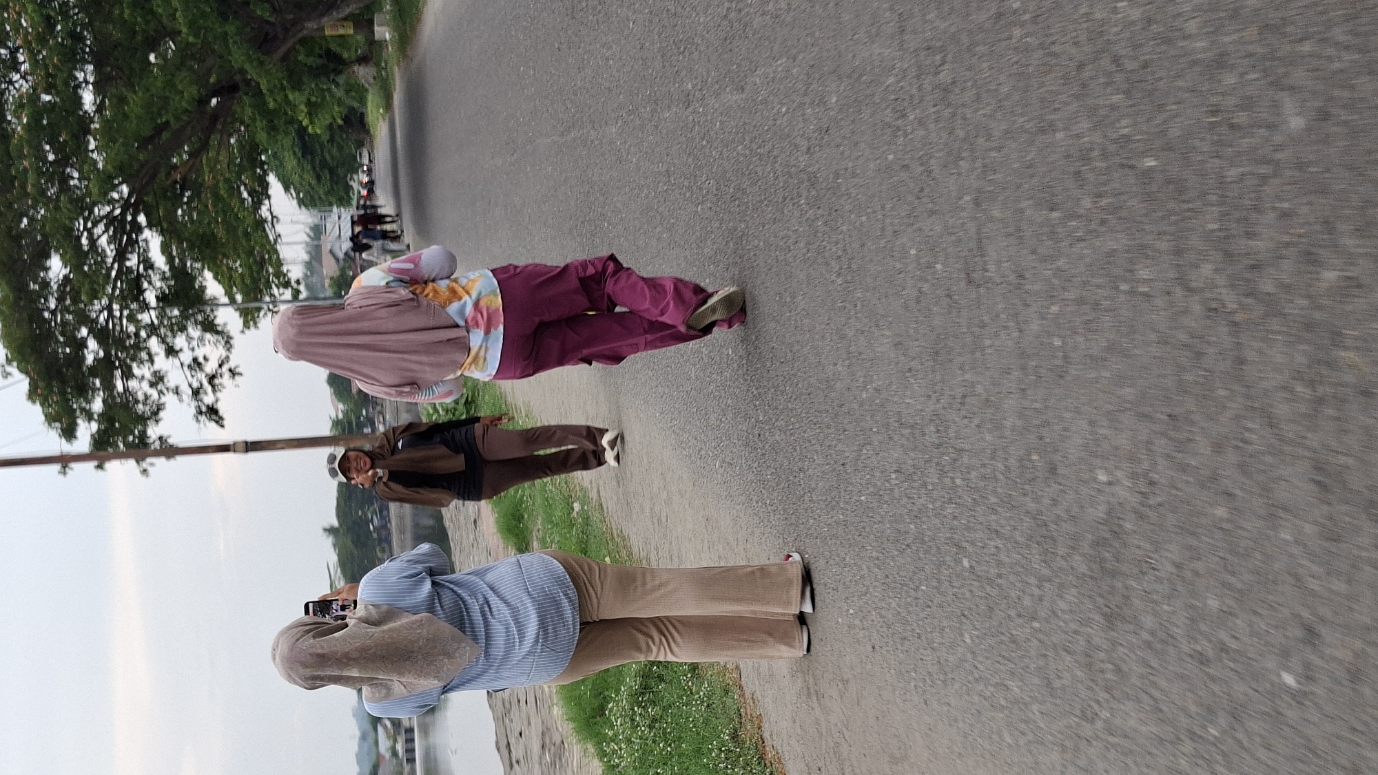 In Lhokseumawe, Indonesia, three women are walking on a paved road next to a body of water. The woman in the foreground on the left, facing away from the camera, wears a light blue pinstripe shirt and light brown flared pants. She is holding a smartphone up, seemingly taking a picture or video. The woman in the middle, also facing away, wears a dark brown hijab, a dark top, and brown pants. She is looking back and smiling at the camera, with one hand raised and fingers pointed towards the camera. The woman on the right, furthest from the camera, faces away from the viewer and wears a pink hijab, a colorful patterned long-sleeved shirt, and dark pink cargo pants. Behind them, the road continues, lined with trees on the right side. In the background, several other people are walking, and there are some buildings and structures visible across the water. The sky is overcast.