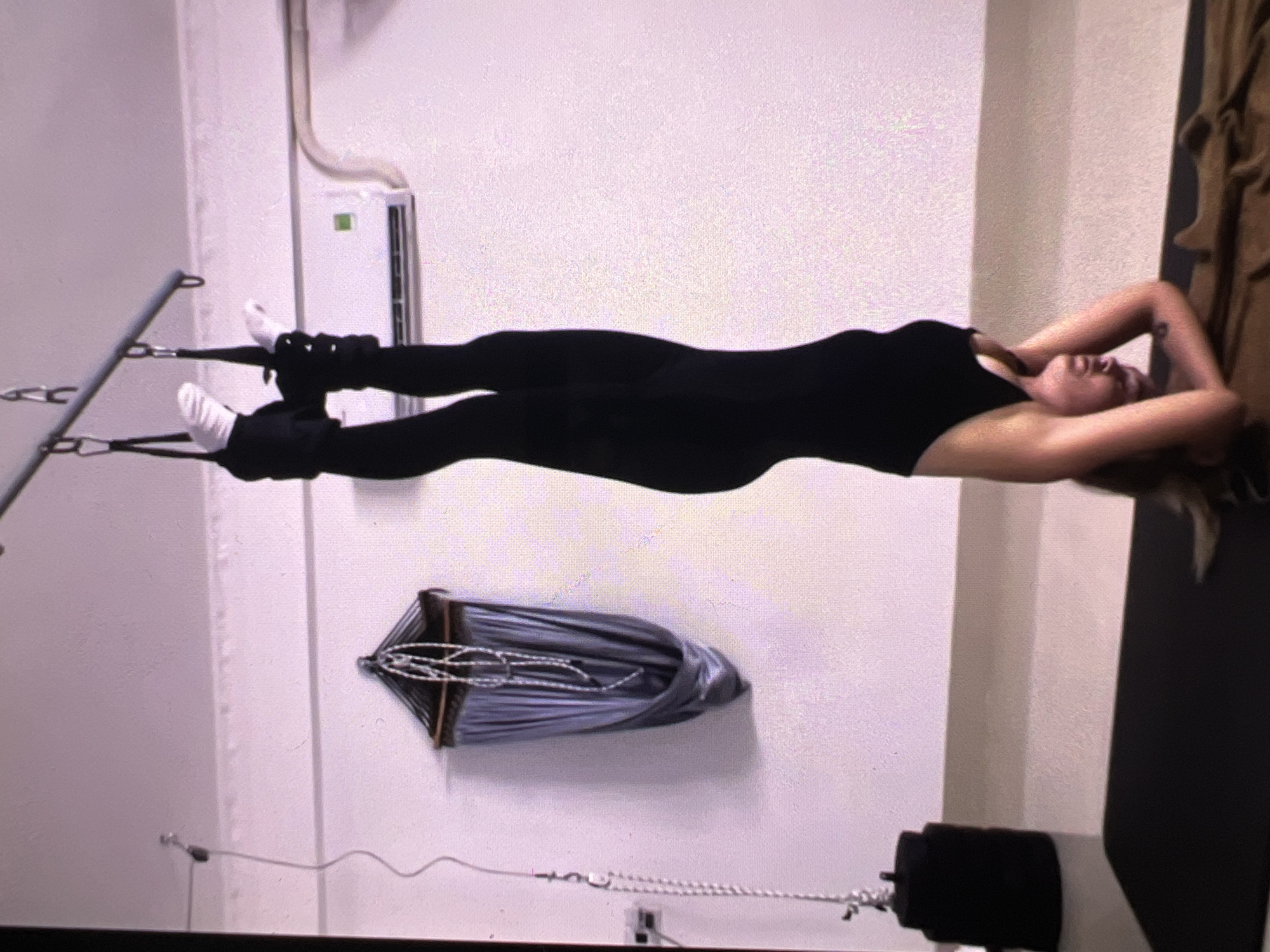 An indoor scene depicts a woman engaged in a fitness exercise, likely Pilates or aerial yoga. She is lying supine, suspended horizontally in the air, with her body parallel to the floor. She wears a black, form-fitting jumpsuit or leotard and white socks. Her feet are secured in black ankle cuffs that are attached by straps to a horizontal metal bar positioned above her head. Her arms are bent, and her hands are clasped behind her head, with her gaze directed upwards.
The background consists of a plain white wall, on which an air conditioning unit is mounted. Below the woman and slightly to her left, a grey, folded fabric, resembling an aerial silk hammock, is suspended from the ceiling. On the far right, a dark, cylindrical object, possibly a weight or a resistance mechanism, rests on the floor with a chain extending from it. A textured brown curtain or fabric is partially visible along the right edge of the frame. The lighting suggests a well-illuminated indoor environment. No text is visible in the image.