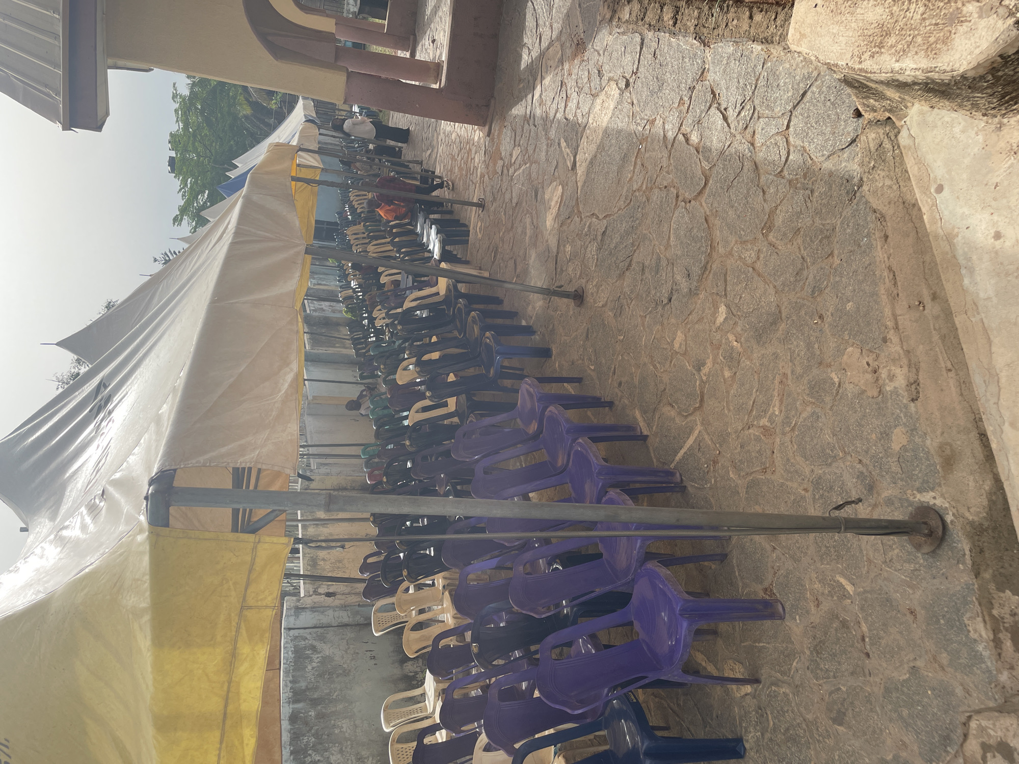 The image depicts a row of plastic chairs under a tent in Akwanga, Nigeria. The chairs are various colors, including white, purple, black, and green. The tent is supported by metal poles. The ground is paved with stone. In the background, there are people visible.