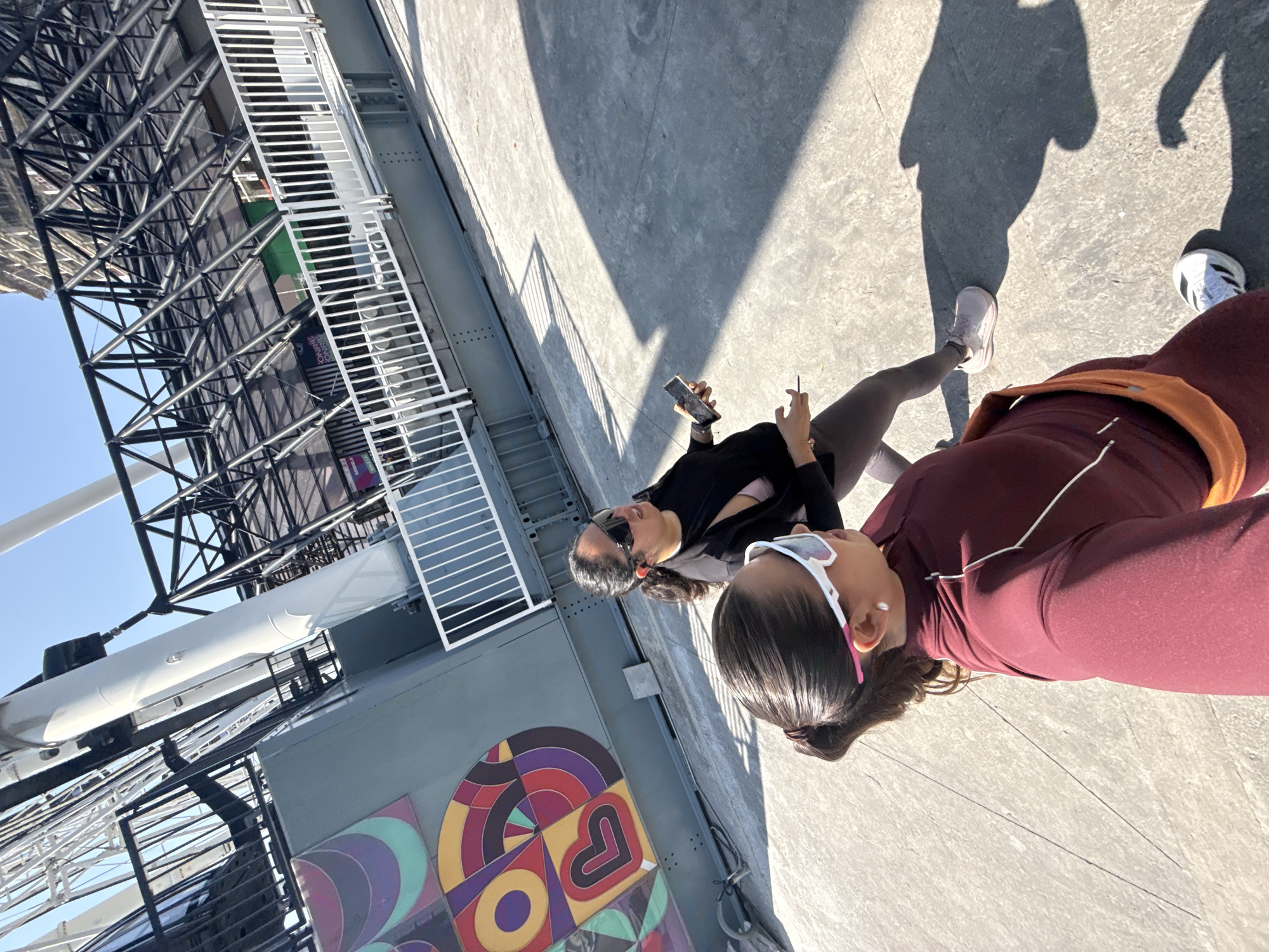The image shows two individuals on a rooftop platform in Puebla, Mexico. One person is wearing a burgundy tracksuit with an orange belt or fanny pack, and white athletic shoes are visible. The other individual is wearing a black top, grey leggings, and light-colored athletic shoes. Both are wearing sunglasses, and one appears to be holding a phone.
The rooftop has a gray concrete surface with shadows indicating a sunny day. Adjacent to the platform is a structure with a colorful, geometric mural featuring shapes such as hearts, circles, and triangles. The platform overlooks a complex metal structure, likely part of a building or architectural feature, with a background of a clear blue sky. The presence of metal scaffolding suggests ongoing construction or maintenance work.