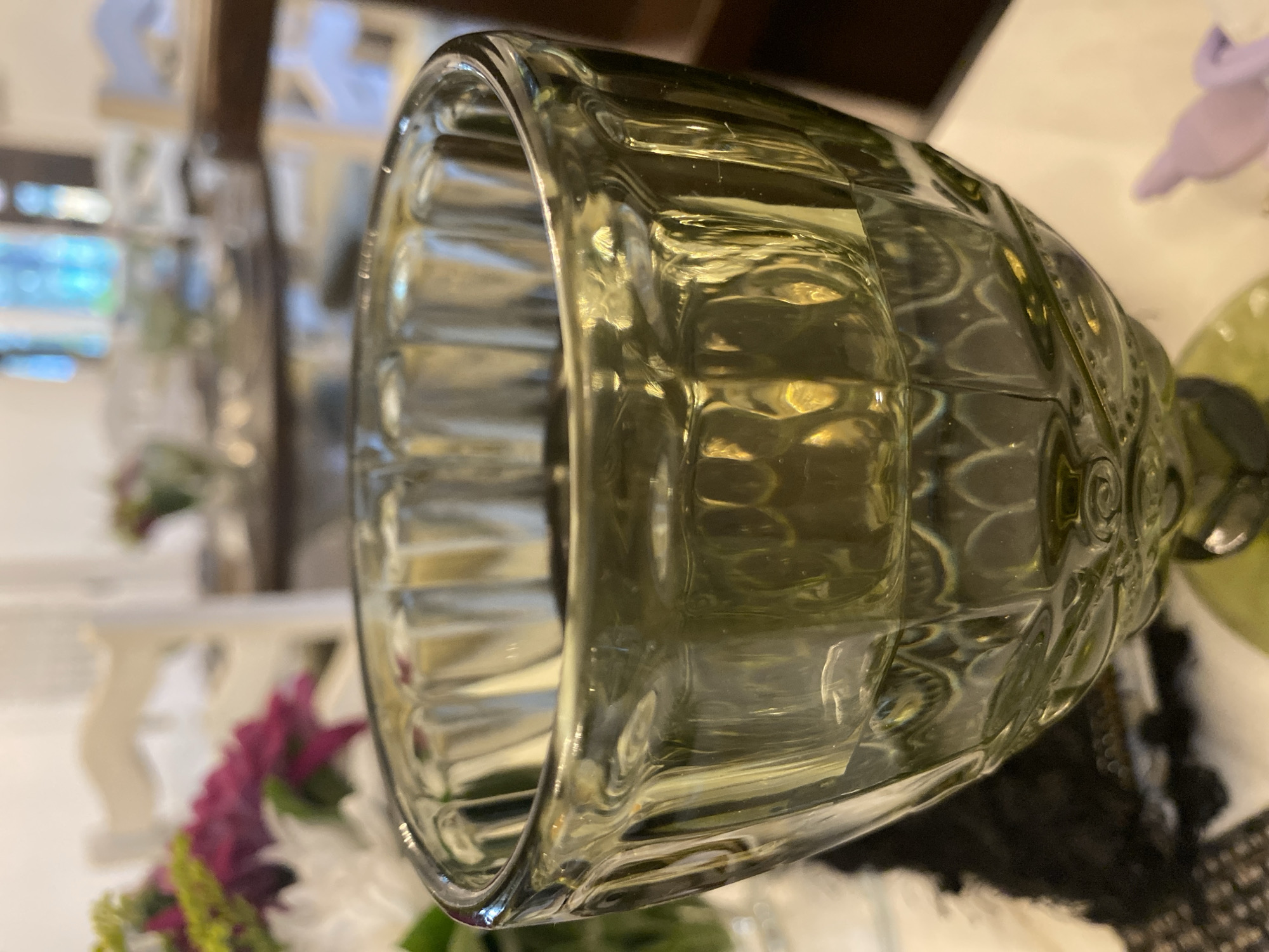 A close-up, slightly tilted shot focuses on a green, glass votive holder filled with liquid and resting on a white tablecloth. The votive holder has a textured pattern of vertical ribs and circular indentations. In the blurred background, a bouquet of white, pink, and green flowers is visible, alongside the back of a white chair. Further in the background, more chairs and tables suggest a dining or event setting. The lighting is soft and diffused, creating highlights and reflections on the glass. The scene is indoors. There are no people visible in the image. The provided information about Davao City, Philippines, is not visually represented in the image and cannot be determined from the image itself.