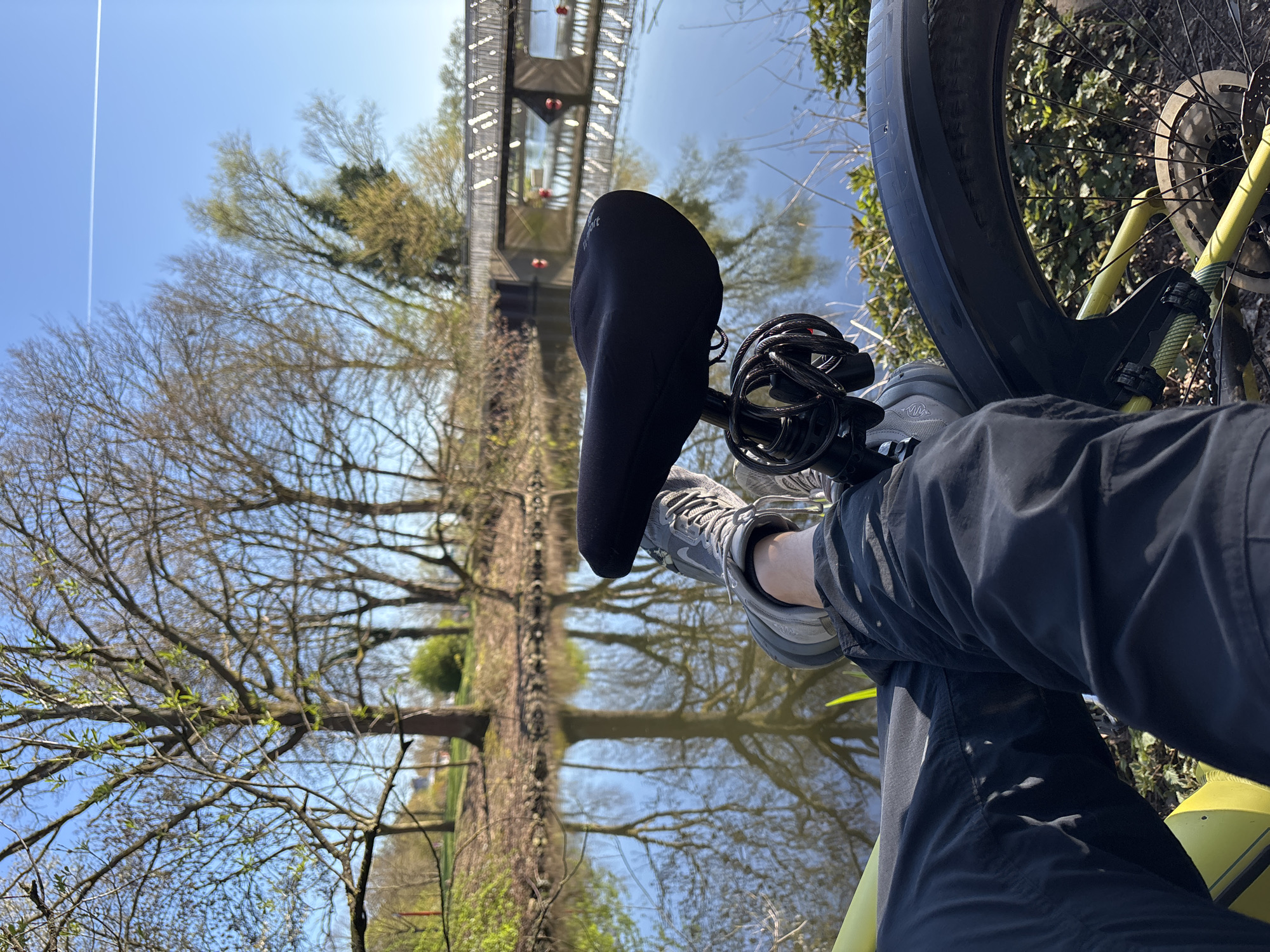 The image shows a cyclist's perspective while stopped. A person wearing gray shoes, black pants, and a black bike shoe cover is seated on a bicycle with a yellow frame. The bike lock is coiled near the seat. The background features a reflection of trees and a bridge in water. The sky is visible in the upper left corner with a contrail. It's likely a warm day as the trees are beginning to get leaves. The location is near a body of water with a pedestrian bridge, but the city and exact location are unknown.
