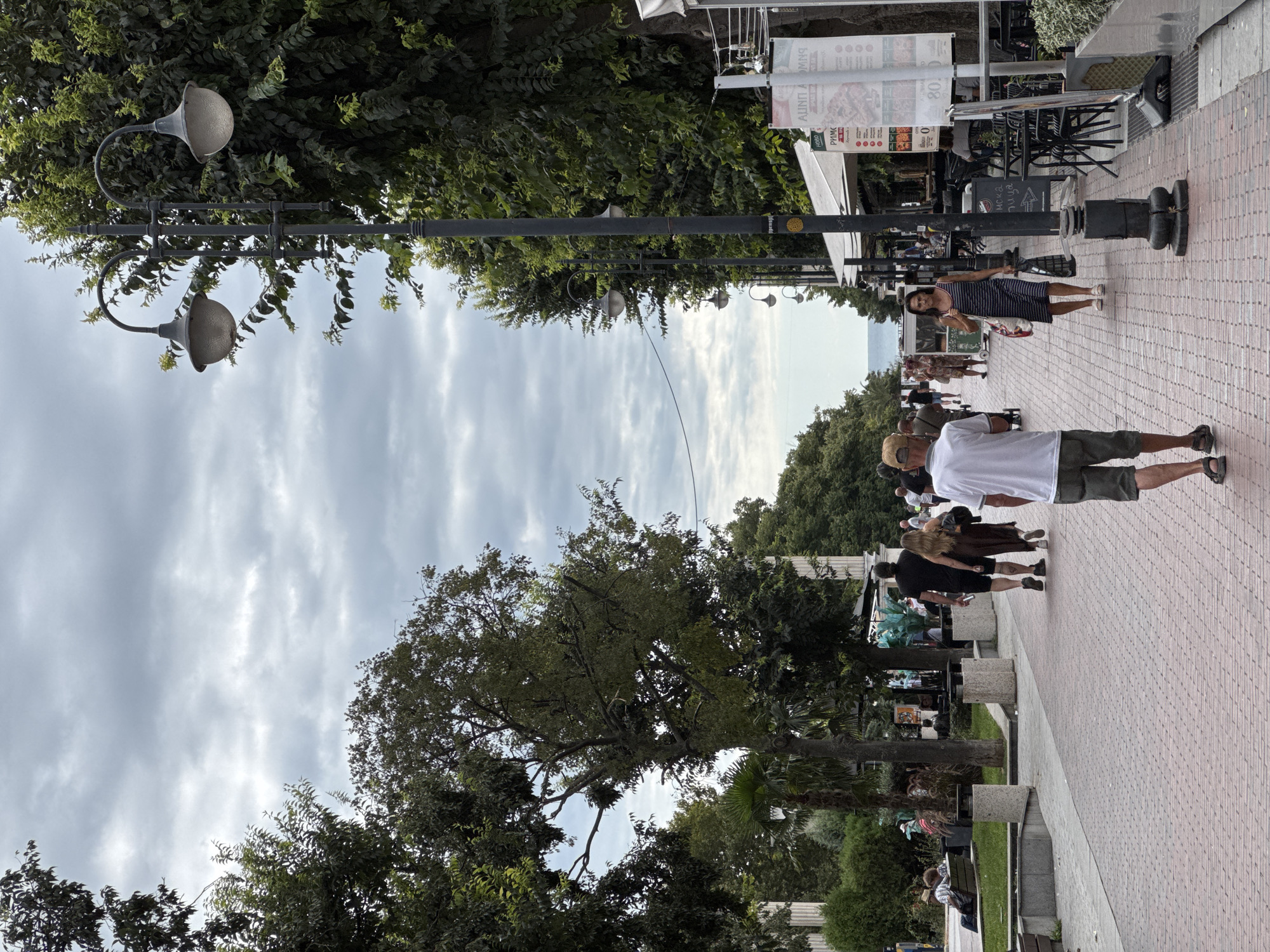 The image depicts a bustling pedestrian promenade in Varna, Bulgaria, likely along the Sea Garden or a similar seaside boulevard, under an overcast sky. A wide, brick-paved walkway is lined with mature trees on both sides, creating a shaded and pleasant atmosphere. Lampposts with ornate, curved arms and globe-shaped lights are spaced along the promenade.
In the foreground, a man wearing a white t-shirt and olive-green shorts walks away from the camera. To his left, a couple walks hand-in-hand, the woman in a dark brown dress and the man in a black t-shirt and shorts. Further down the promenade, several other individuals are visible, some walking, others seated on benches or at outdoor cafe tables. A woman in a striped dress walks towards the camera carrying shopping bags.
On the right side of the promenade, outdoor seating areas for cafes and restaurants are set up. Signs, some with Cyrillic script, advertise services. The vegetation is lush, with a variety of trees and plants contributing to the green environment. In the background, beyond the trees and the promenade, a glimpse of the Black Sea is visible under the cloudy sky. The overall impression is one of a lively public space in a coastal city.