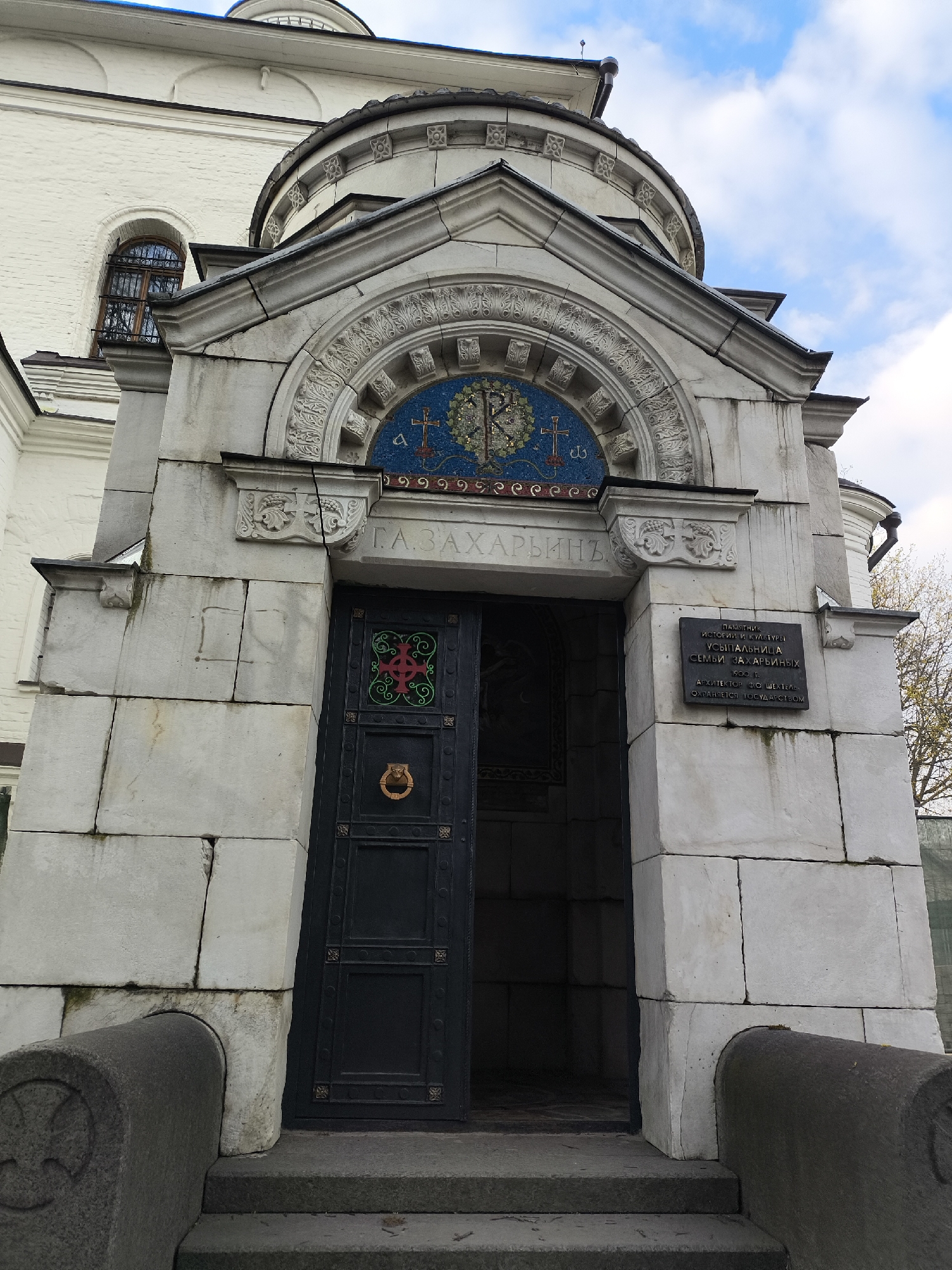 The image shows the entrance to the Zacharin family vault, located in Moscow, Russia. The entrance is made of gray stone blocks and features a dark green door that is partially open. Above the door is a mosaic depicting a cross and floral motifs. A plaque on the right side of the entrance indicates that the vault is a monument of history and culture, dating back to 1900 and designed by architect F.O. Schechtel. The inscription above the door reads "Г.А.ЗАХАРЬИНЪ" (G.A. Zakharin). Stone steps lead up to the entrance, and a section of a white building is visible in the background. The sky is partly cloudy.