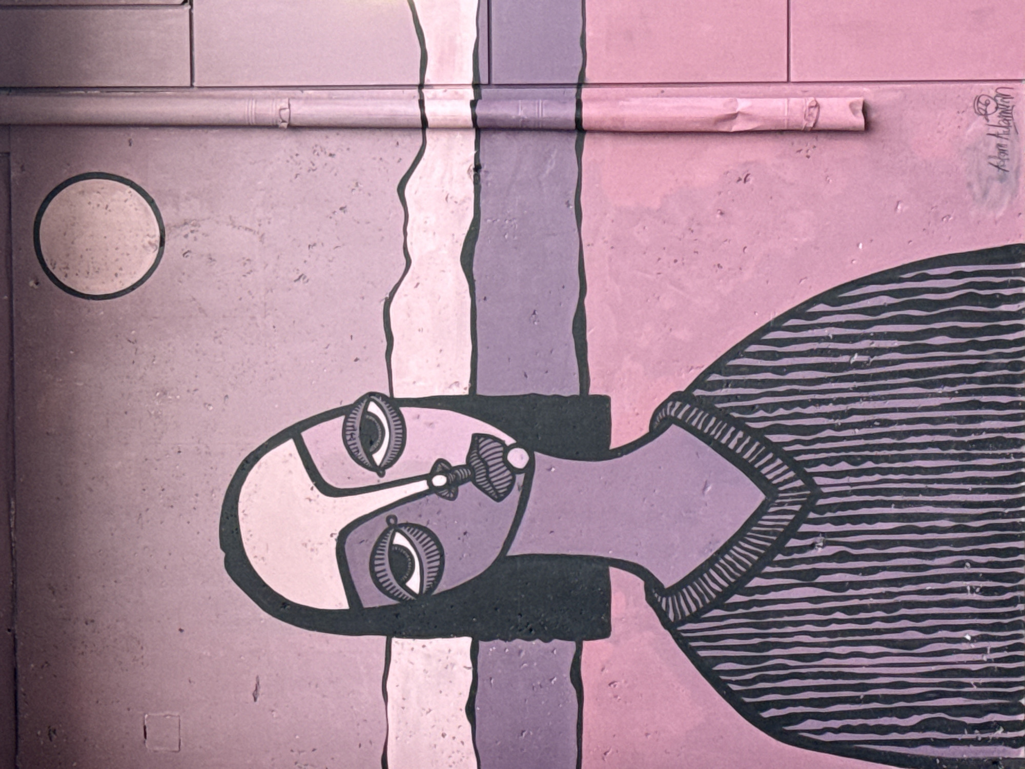 This is a close-up of a mural or street art in Yerevan, Armenia, featuring a stylized human figure. The artwork is painted on what appears to be a wall or door composed of panels. The color palette is predominantly shades of pink, lavender, and gray, with black accents.
The figure is depicted in a somewhat abstract or cubist style, with oversized eyes, a bulbous nose, and a simplified body. It has a striped garment on. To the left of the figure is a small circle.
Above the artwork runs a horizontal cylindrical object, possibly a pipe, which is painted the same pinkish-lavender color as the background.
In the upper right corner of the image is a signature or mark "Adam Adamyan". The mural is marked with signs of weathering or age, adding to the urban aesthetic.