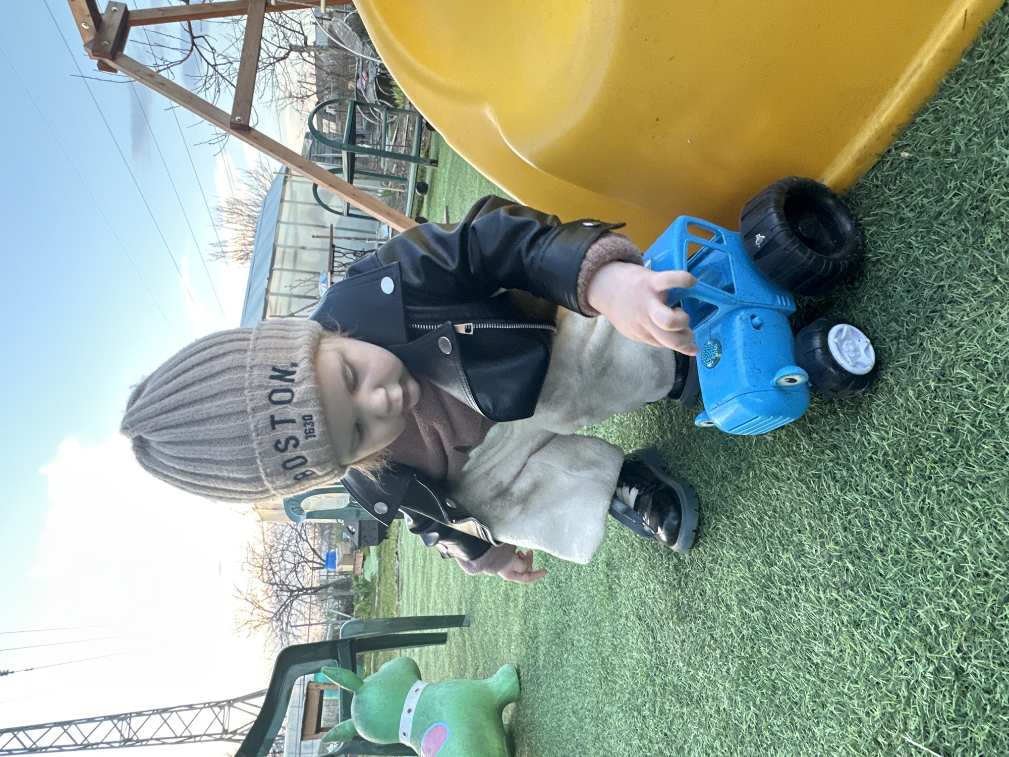 A child in Chaikovsky, Russia, wearing a tan knit hat with the word "BOSTON" knitted on it, a black leather jacket, and beige pants, is kneeling on artificial turf next to a yellow plastic slide. The child is touching a blue plastic toy truck with black and white wheels. A green plastic toy animal is visible near the child, and a green metal chair is behind it. A metal playground structure is in the background with a greenhouse visible behind the structure. The sky is light blue.