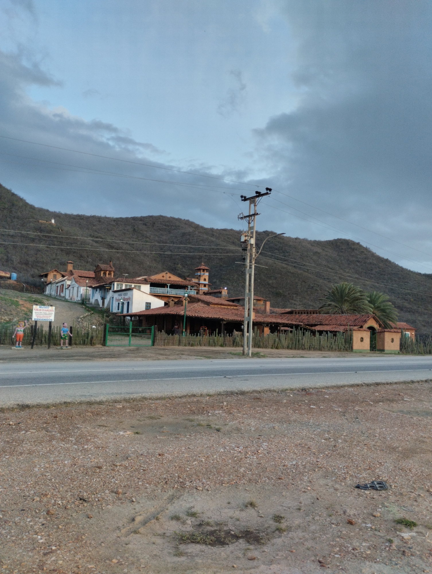 The image depicts a roadside scene in Juan Griego, Venezuela, featuring a collection of buildings with terracotta roofs nestled against a barren, mountainous backdrop. A paved road runs horizontally across the foreground, with a gravel shoulder bordering it. To the right of the road, a utility pole with electrical equipment stands prominently.
The buildings appear to be part of a cultural or historical site, possibly a museum, as indicated by a sign visible in the cropped image that reads "MUSEO PUEBLO DE MARGARITA" and "BIENVENIDOS." The architecture suggests a traditional Spanish colonial style, with white stucco walls, arched doorways, and exposed wooden beams. Some of the buildings have multiple stories, and one features a tower with a distinct architectural design.
A low, green metal fence separates the road from the property, and a line of cactus plants forms a natural barrier along the fence. Two human figures are visible near the entrance of the property, one standing by the sign and another to its right. The landscape is arid and dry, with sparse vegetation on the slopes of the mountains. The sky is overcast with dramatic clouds, suggesting a change in weather. The overall impression is one of a remote, historical destination situated in a rugged, natural environment.