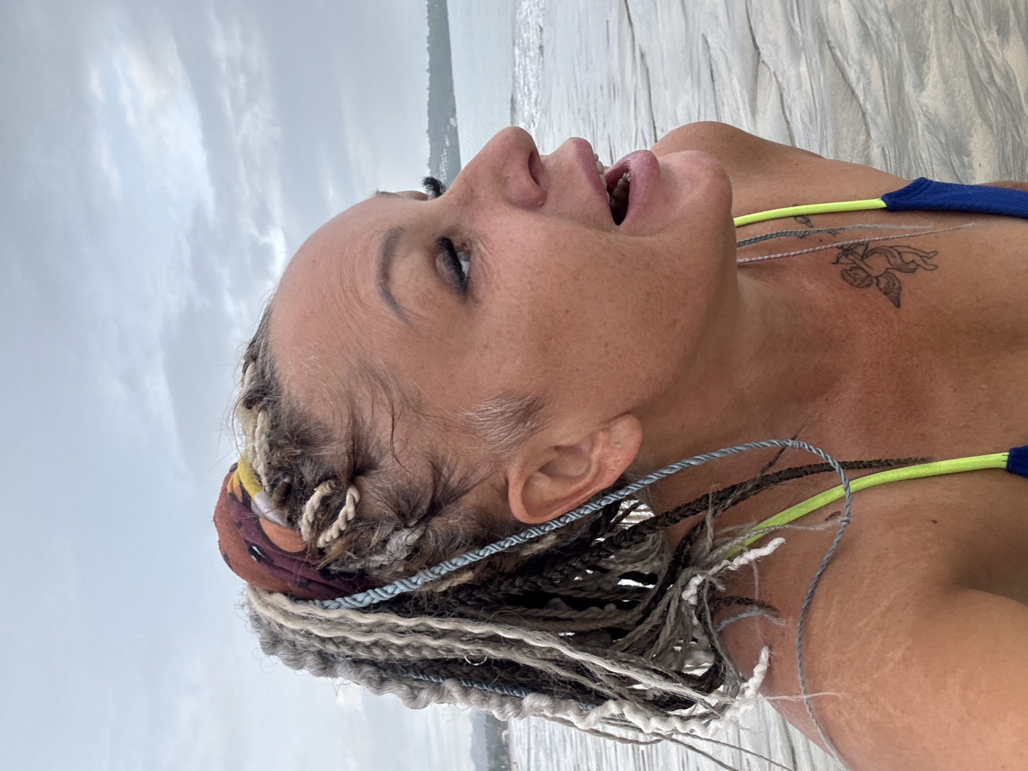 The image shows a woman with braids looking up at the sky on a beach in Kuta Selatan, Indonesia. She has a tattoo on her chest. The sky is cloudy, and the ocean is visible in the background.