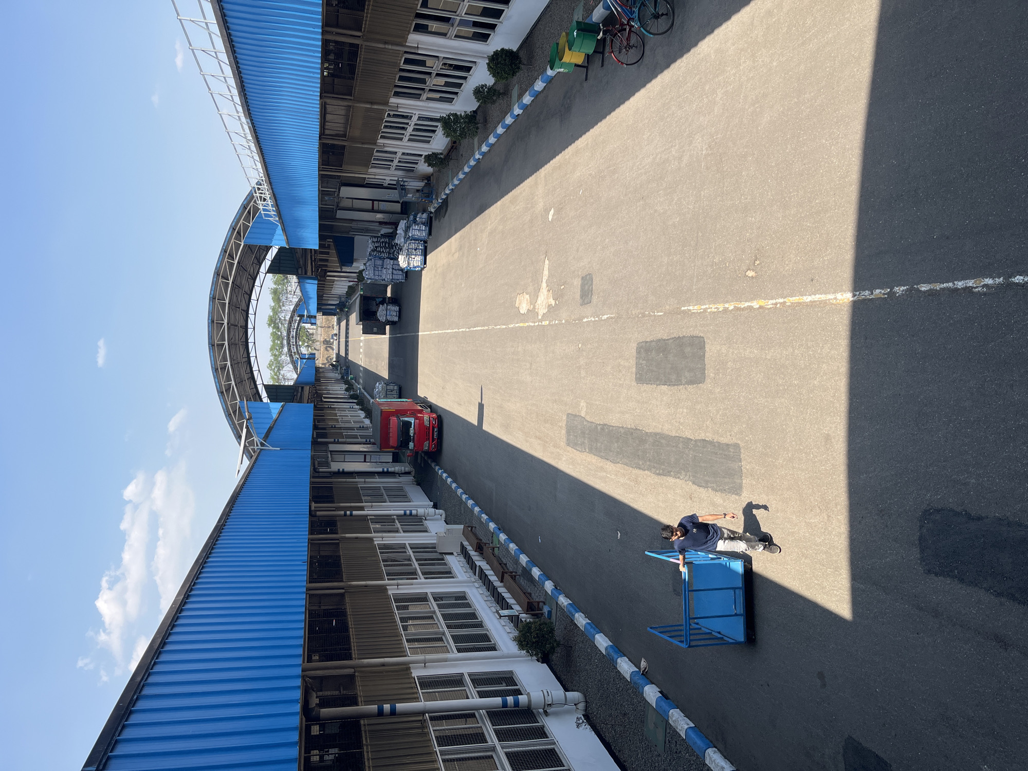The image depicts a wide, paved outdoor area between two long industrial buildings with blue corrugated metal roofs supported by an arched, open-air structure. The buildings have white facades with numerous regularly spaced windows and dark, possibly metal, cladding at the lower level. A blue and white striped curb runs along the edges of the paved area.
In the foreground, a young man wearing a dark blue t-shirt and light-colored pants is pushing a bright blue metal cart. He is walking on the paved surface, casting a shadow.
Further into the scene, a red truck is parked, and stacks of white bags or bales are visible on pallets, some of which are on trolleys. Bicycles and green trash bins are situated near the right-hand building. The sky is clear and blue with a few scattered white clouds. The overall environment suggests a logistics or manufacturing facility, potentially located within an industrial zone in Pelang, Indonesia, given the context provided.