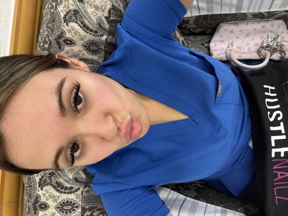 A woman wearing bright blue scrubs with a "FIGS" tag is lying on a patterned pillow. She has long eyelashes and pursed lips. Next to her is a small, pink floral purse with a handle and a silver charm. A black bag with the white text "HUSTLENAILL" is partially visible. The image is taken from a high angle.