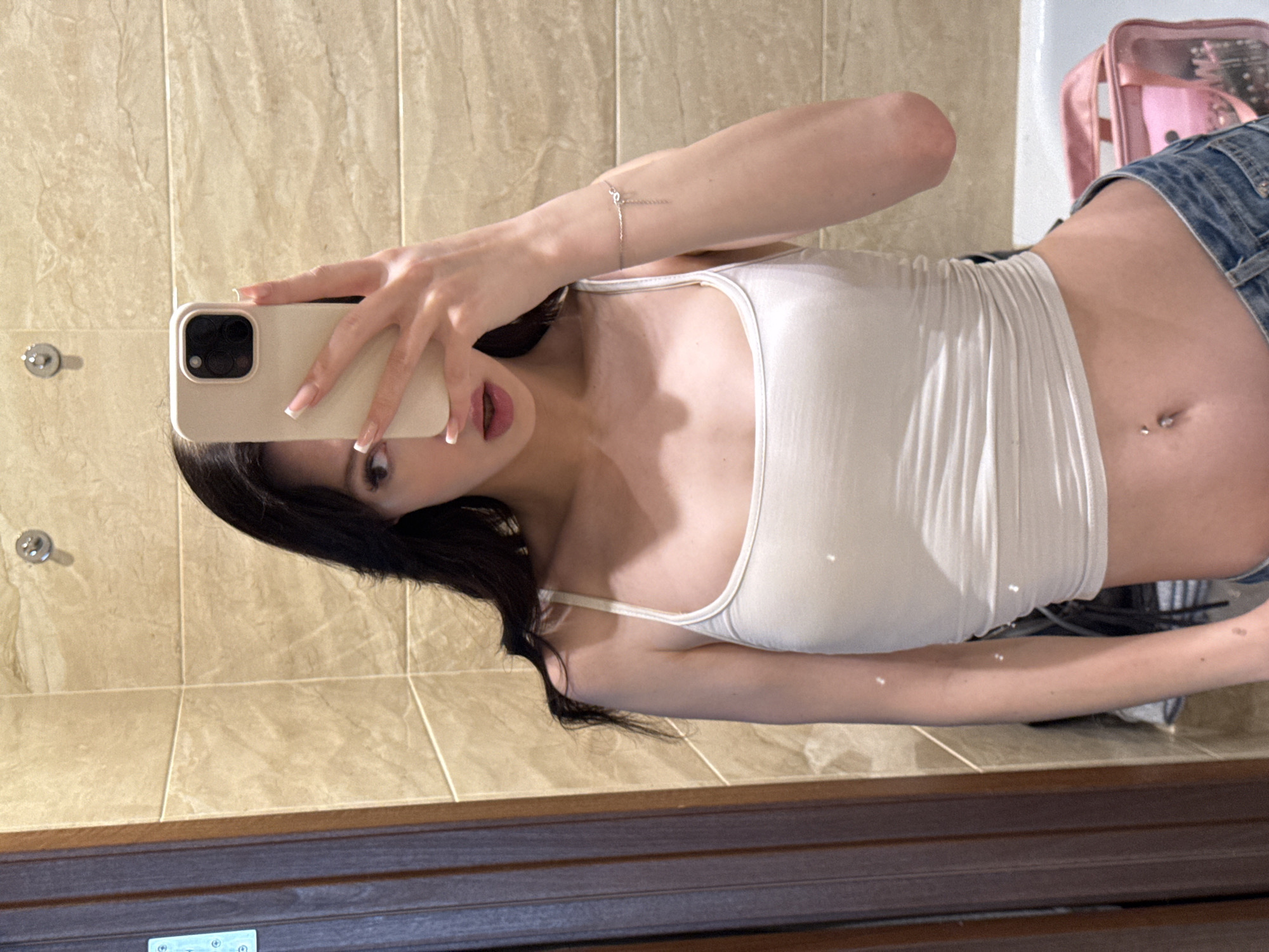 A young woman is taking a mirror selfie in what appears to be a bathroom with beige tiled walls. She is wearing a white tank top and denim shorts. Her hair is dark and long, and she is holding a light-colored phone in her right hand to take the picture. She has a bracelet on her left wrist and a belly button piercing. In the background, a pink toiletry bag is visible. The lighting suggests it is daytime. The overall mood seems casual and playful. No visible text.