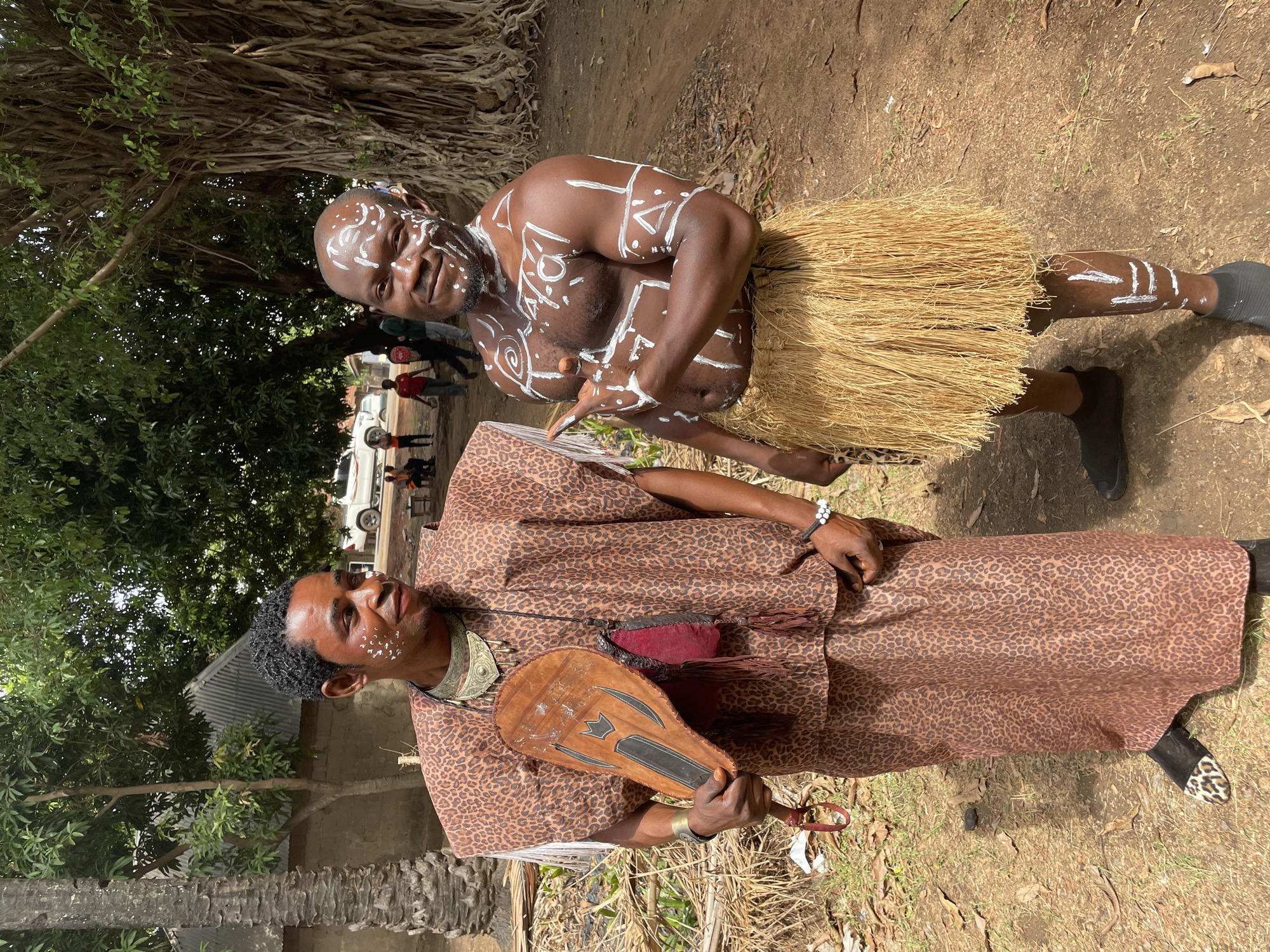 Two men stand outdoors on a patch of dry ground. The man in the foreground wears a brown leopard-print patterned outfit with a decorative necklace, holding a wooden fan-like object. The man behind him is bare-chested with white body paint and a straw skirt, wearing dark socks. The background contains greenery, a building, and parked cars. The location is Akwanga, Nigeria.