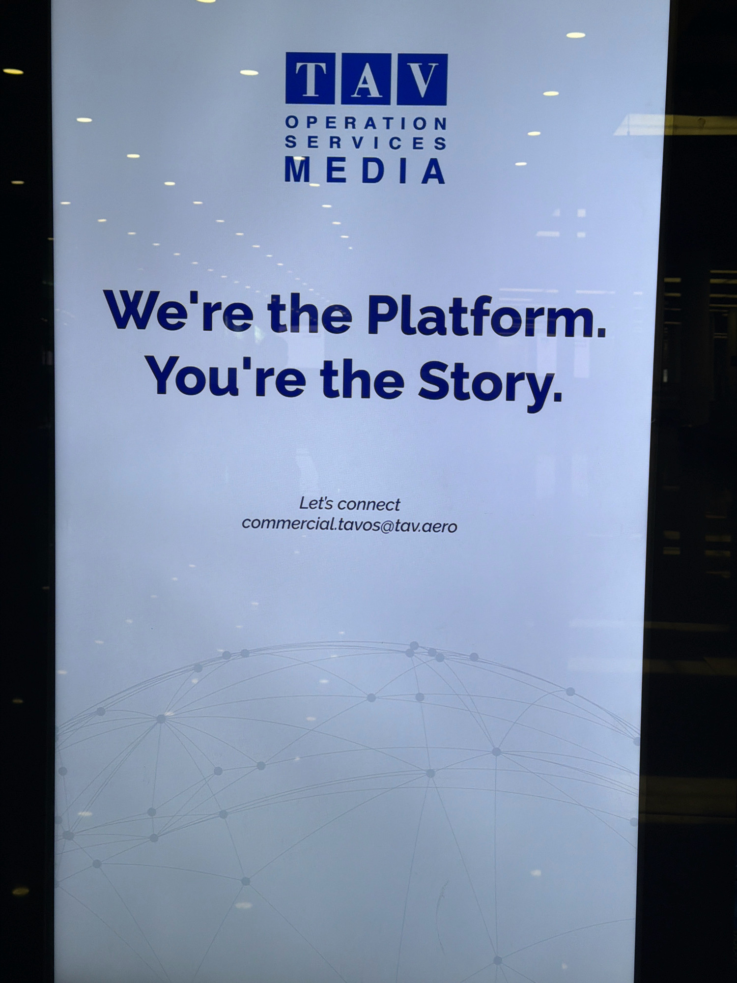 The image shows a brightly lit vertical banner with text and a logo. At the top, a blue logo contains the letters "TAV" within squares. Below the logo, the words "OPERATION SERVICES MEDIA" are printed in capital letters.
The main text on the banner reads: "We're the Platform. You're the Story." This text is in a large, dark blue, sans-serif font.
Below this, smaller text says: "Let's connect commercial.tavos@tav.aero".
The background of the banner is a light blue-grey, and at the bottom, there is a faint graphic of a network of interconnected dots and lines, resembling a globe or a web. The banner appears to be illuminated from the front, and there are reflections of overhead lights and possibly the surrounding environment on its surface.
The image quality is high, with sharp focus on the text and logo. The overall impression is of a professional advertisement or informational display. There are no people visible in the image. The banner is positioned in what appears to be an indoor setting, possibly a commercial space or an airport terminal, given the nature of the advertisement. The context of Gaziemir, Turkey, is noted, suggesting this banner is located within or related to facilities in that region, such as the Izmir Adnan Menderes Airport, which is served by TAV Airports.