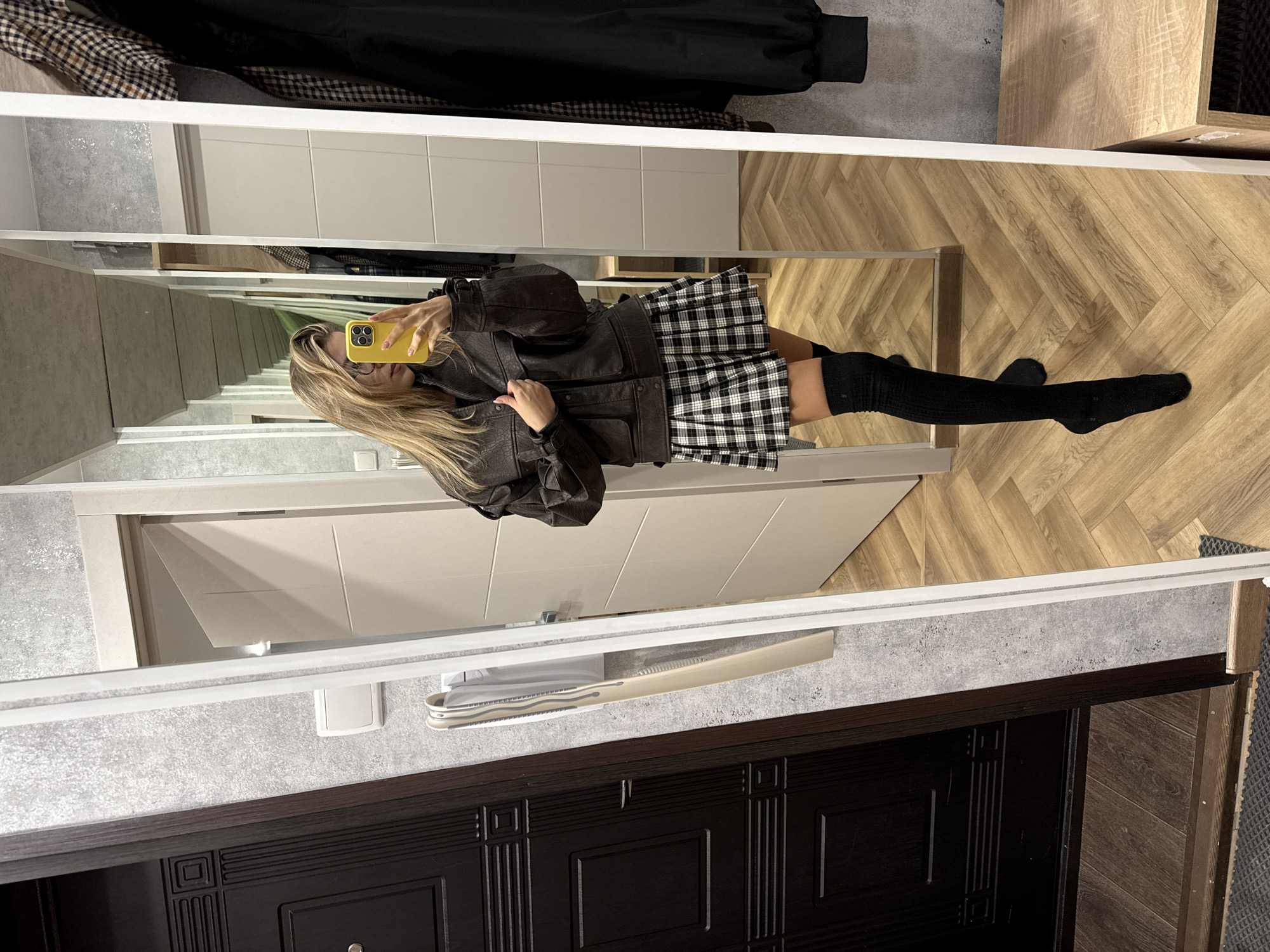 A full-length mirror selfie, rotated 90 degrees clockwise, depicts a person in an indoor hallway or entryway. The individual, with long blonde hair and wearing glasses, holds a bright yellow smartphone to capture their reflection. They are dressed in a dark brown or black bomber-style jacket, a black and white plaid pleated mini-skirt, and black over-the-knee socks.
The mirror's reflection shows a light-colored wooden floor with a herringbone pattern and white panel doors or cabinets in the background. Clothes, including some with a checkered pattern, are partially visible hanging on a rack within the reflection.
In the foreground, outside the mirror's frame, is a dark brown or black wooden door featuring decorative paneling. The floor beneath the mirror is a light gray, possibly concrete or tiled surface. A white wall-mounted object, likely a light switch, and a long, thin white object (possibly a shoe horn) are visible on the wall. To the right of the dark door, a section of dark wooden flooring with a herringbone pattern and a dark mat or rug are present. The setting is brightly lit, consistent with an interior residential space. The time of day and external weather conditions are not evident. No distinct text is visible.
