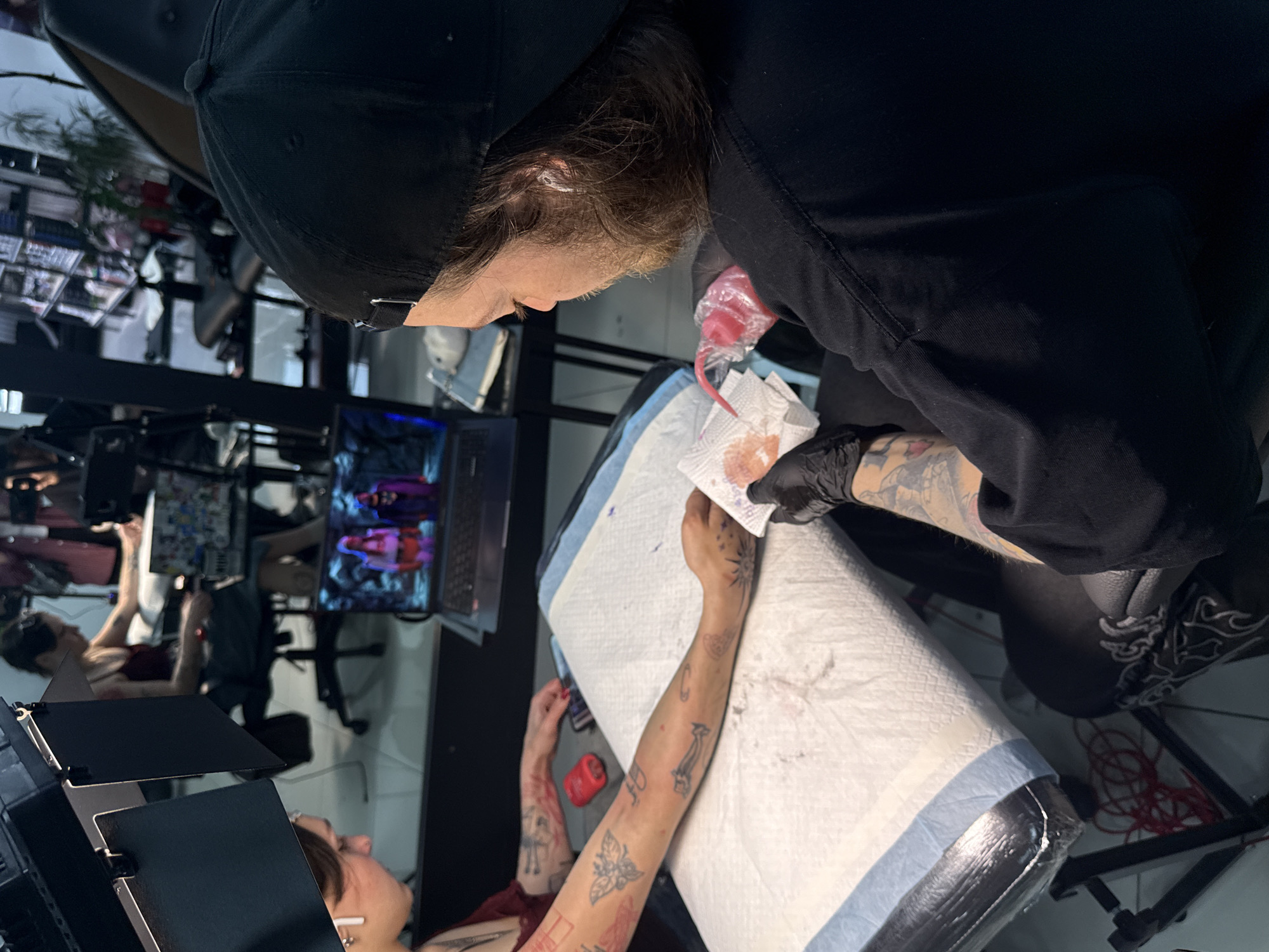 A tattoo artist wearing a black cap and black gloves is working on a client's arm in a tattoo studio. The client's arm is covered in tattoos and is lying on a white and blue padded surface. The artist is using a tattoo machine with a red ink bottle attached. A bloody paper towel is next to the tattoo being performed. In the background, there is a laptop and monitors with various images displayed on them. In the distance, there is another person being tattooed. The location appears to be a professional tattoo studio, potentially in Rostov-on-Don, Russia.