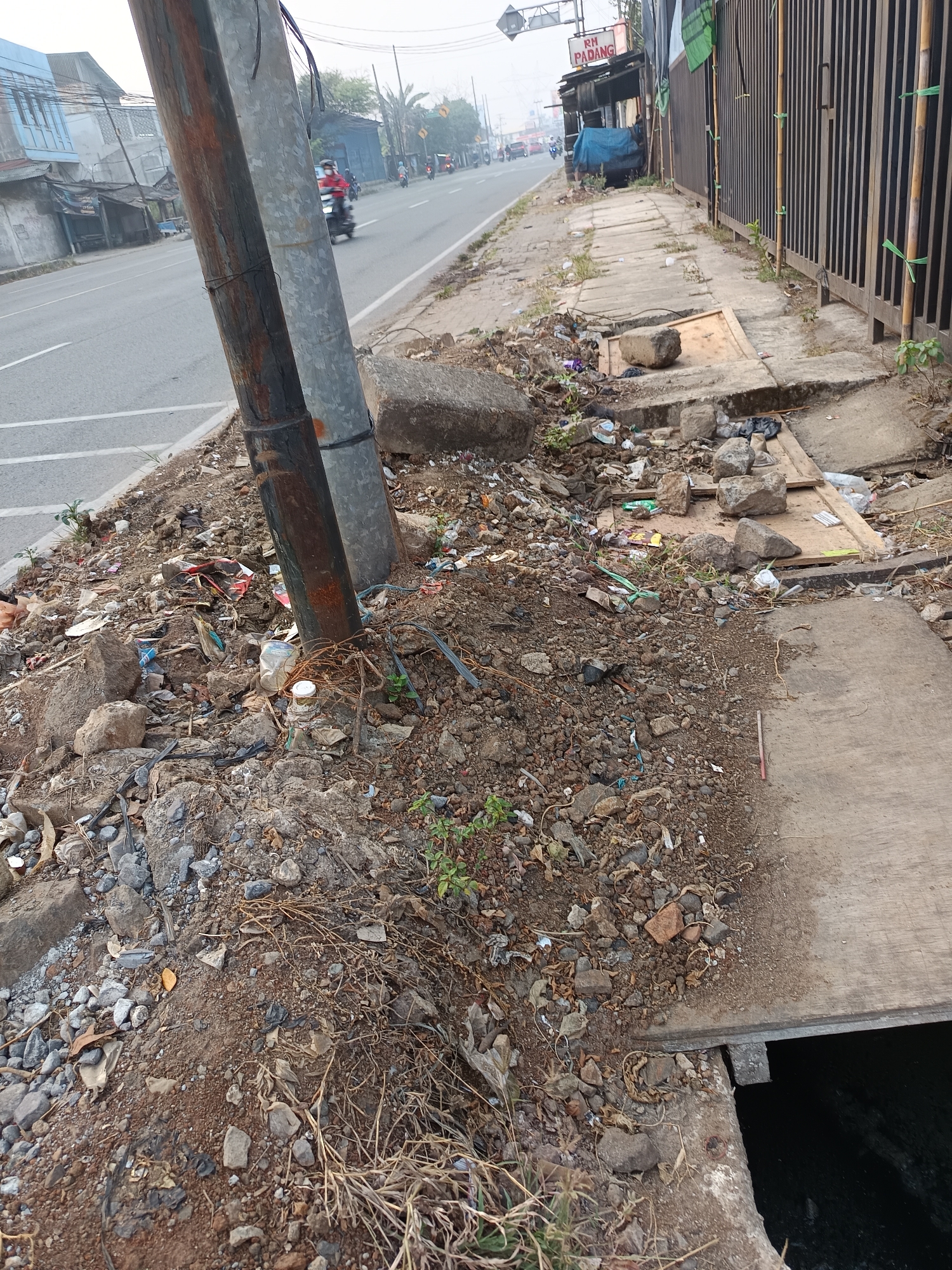 The image depicts a cluttered roadside in Talaga Sari, Indonesia. A pole, likely for utility lines, stands prominently in the foreground, its base surrounded by debris. The roadside is littered with rocks, soil, dried vegetation, and various discarded items, including plastic bags, bottles, and wrappers. A concrete sidewalk runs parallel to the road, also showing signs of disrepair and accumulation of trash. Beyond the sidewalk, a metal fence encloses an area. The road itself is paved, with white lane markings visible. Several motorcycles are in motion on the road, indicating active traffic. Buildings and structures line the opposite side of the road, with one bearing a sign that reads "RH PADANG". The overall environment appears to be an urban or semi-urban setting characterized by neglect and the presence of waste.