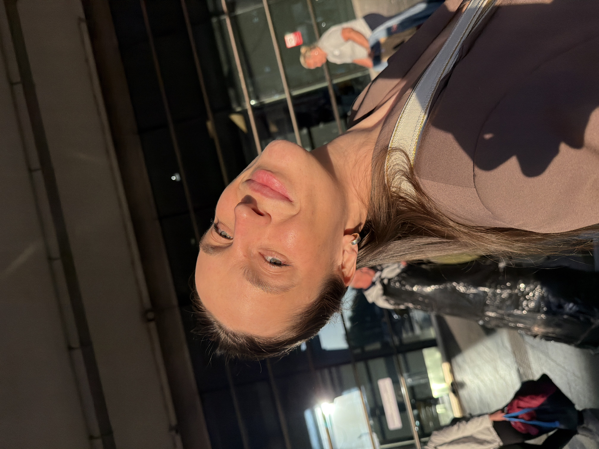 A medium shot presents a woman with light skin, facing the camera at an angle, against an urban background. She has light-colored eyes, pink lips, and light brown hair. She is wearing a brown jacket with a light-colored strap running diagonally across her shoulder. An earring is visible on her left ear. The background shows a modern building with glass windows and a metallic frame. Through the windows, indistinct figures and objects are partially visible, suggesting a bustling city scene. The light indicates that the photo may have been taken in the evening or early morning, as there is a strong contrast of light and shadow. The photo is taken outdoors in the city of Unknown, Unknown.