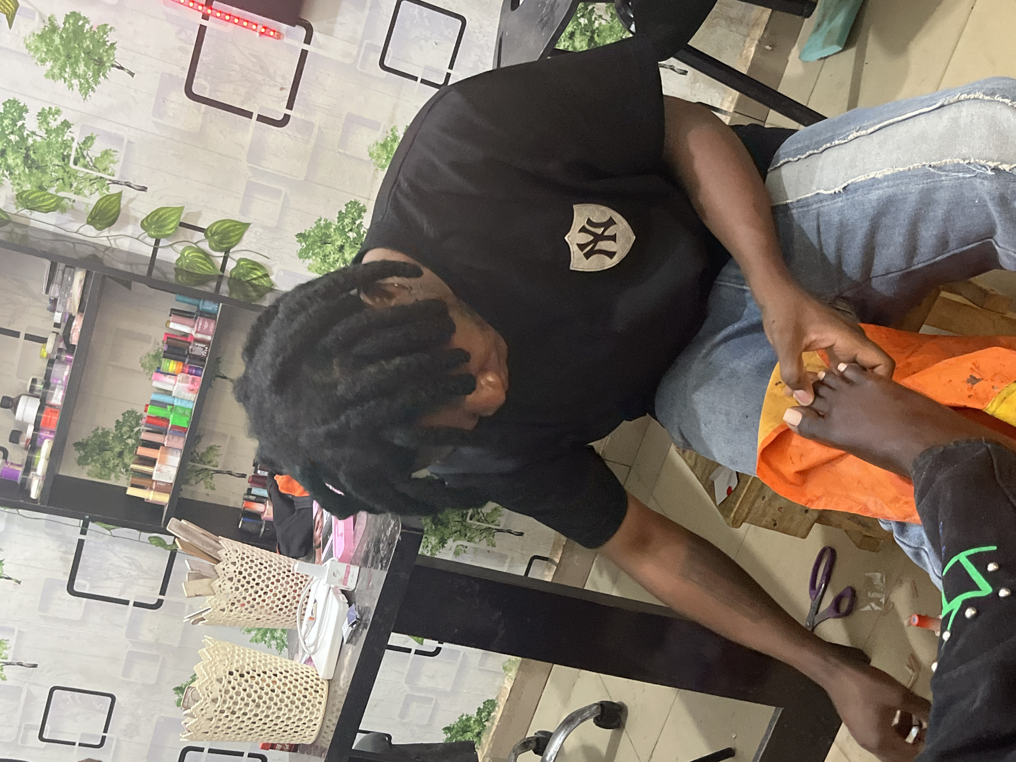 An indoor scene within a commercial establishment, likely a nail or beauty salon, is captured. A dark-skinned individual, the service provider, is seated on a low wooden stool, actively performing a pedicure on the foot of a dark-skinned client.
The service provider is wearing a black short-sleeved t-shirt with a shield-shaped "NY" emblem on the left chest and blue jeans. They have dark dreadlocks and are leaning forward, concentrating on the client's right foot, which they hold with both hands. The client's lower right leg and foot are visible, resting on an orange fabric. The client's lower leg is clad in a black garment adorned with green linear designs and silver stud embellishments. Several toenails on the client's foot are painted with a light tan or nude polish.
To the left, a dark-colored table holds two light-colored, perforated containers filled with various tools, including slender wooden sticks. Additionally, a white bottle, a white rectangular electronic device, a small pink box, and small silver packets are arranged on the table surface. A pair of purple-handled scissors lies on the light-colored tiled floor near the service provider's feet.
The background wall is covered with wallpaper featuring a pattern of white squares, darker square outlines, and scattered green tree and foliage motifs. A vine with green leaves hangs vertically down the wall on the left. Black shelving units are mounted on the wall, displaying numerous bottles of nail polish, powders, and other salon products. A vertical strip of red LEDs is visible on the far right of the background. The specific location of Jos, Nigeria, cannot be confirmed through visual cues within the image.
