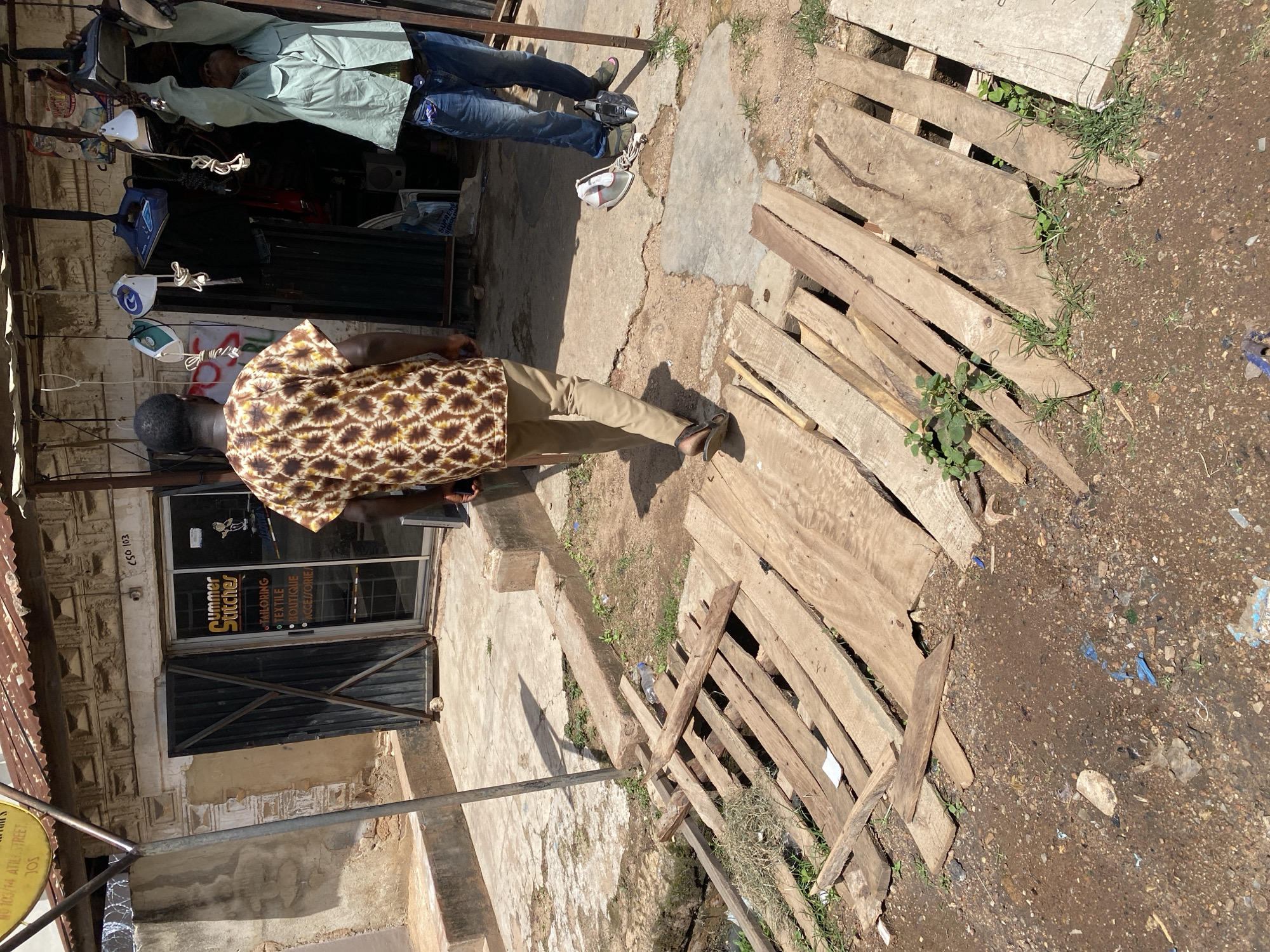 The image depicts an outdoor scene in front of a shop in Jos, Nigeria. A man wearing a brown and yellow patterned shirt and tan trousers is walking away from the camera towards the shop. Another person, wearing a light green shirt and blue jeans, is standing to the right of the shop and holding an object overhead. The shop itself appears to be a tailor's shop, with "Summer Stitches" visible on the signage. Inside, clothing and various items, possibly related to tailoring or repair services, are displayed. In the foreground, a makeshift wooden bridge or walkway is constructed over a ditch or drain. The ground is a mixture of dirt and gravel, with scattered debris and some vegetation growing. The overall environment suggests a bustling commercial area in an urban setting, with signs of everyday life and economic activity.