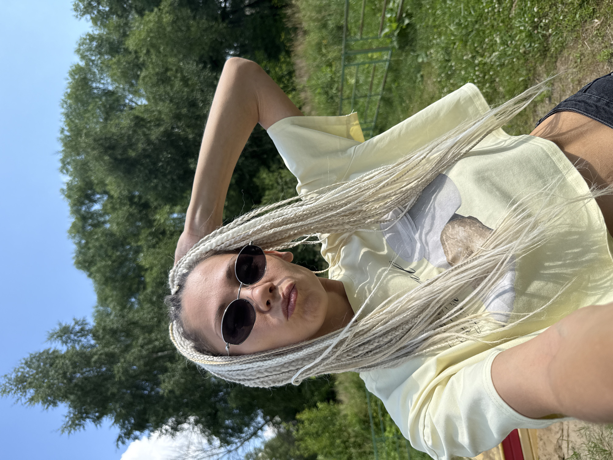 A woman with blonde braids, wearing a light yellow crop top and sunglasses, poses for a selfie outdoors with a backdrop of trees and a blue sky. She is puckering her lips and has her right arm raised, touching her hair. The location is identified as Rusanovo, Russia. The environment appears to be grassy with some fencing visible in the background.
