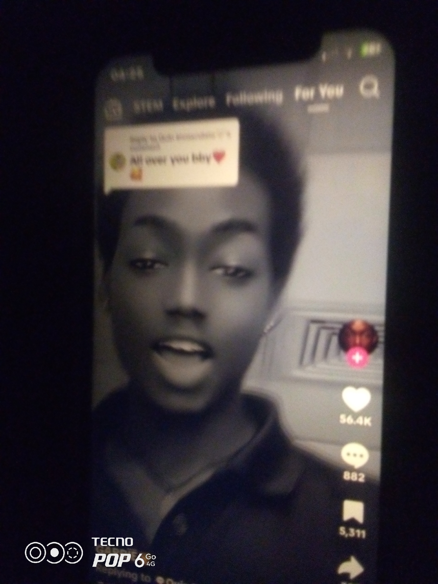The image shows a close-up of a person's face on a smartphone screen, displayed within a social media application. The person, with dark skin and short hair, is looking directly at the camera with an open mouth. The phone screen displays the interface of a video-sharing platform, featuring text overlays such as "All over you bby" with a heart emoji, and interaction icons like a heart, speech bubble, and bookmark, with associated counts of 56.4K, 882, and 5,311 respectively. The top of the screen shows navigation elements with text like "STEM," "Explore," and "For You," and a search icon. The bottom left corner of the image shows the "TECNO POP 6 4G" logo. The overall scene appears to be captured in low-light conditions, suggesting it was taken indoors. There is no indication or visual information within the image that suggests a specific location in Howayi, Nigeria.