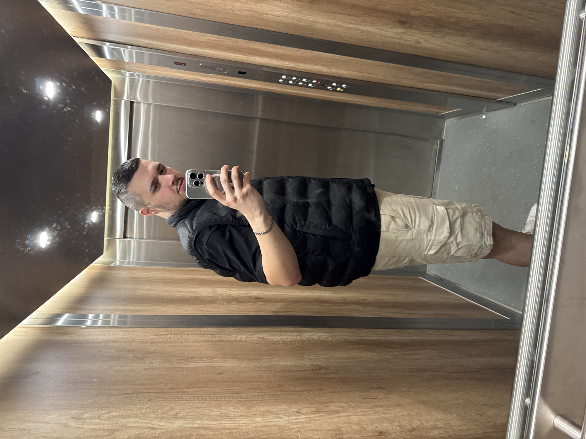 A man is taking a selfie in an elevator. He is wearing a black vest, a black shirt, and khaki shorts. The elevator has wood paneling on the sides and a stainless steel back wall with a panel of buttons. The floor of the elevator is gray. The location is Sofia, Bulgaria.