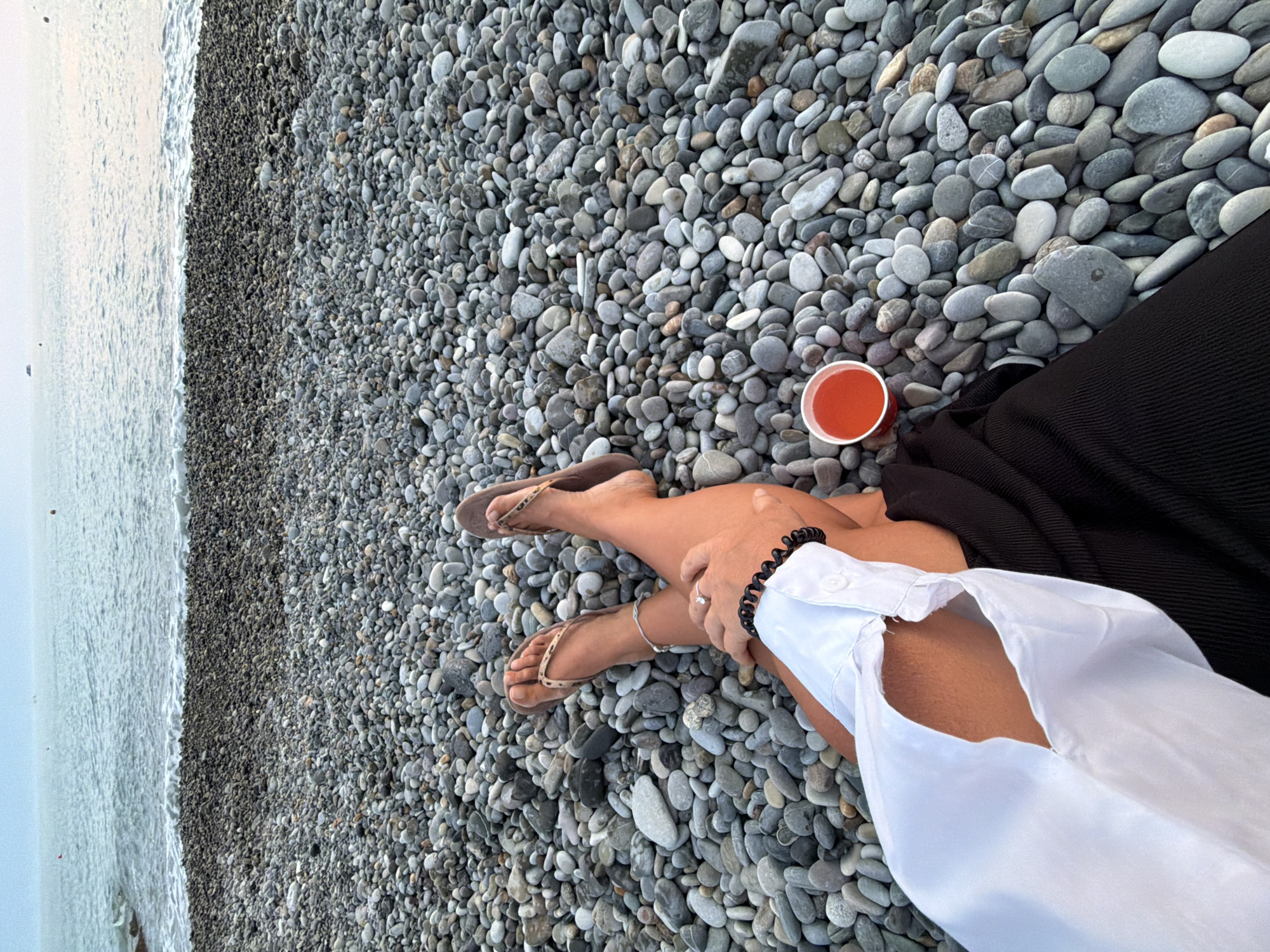 The image shows a person sitting on a pebble beach in Sochi, Russia, facing the sea. The person is wearing a white open-collared shirt and dark bottoms, with their legs crossed. They are wearing flip-flops, a bracelet, and a ring on their left hand. A small cup of red liquid is placed on the pebbles next to them. The sea is visible in the background, with a calm surface and a hazy sky. The beach is covered in a dense layer of grey and white pebbles.