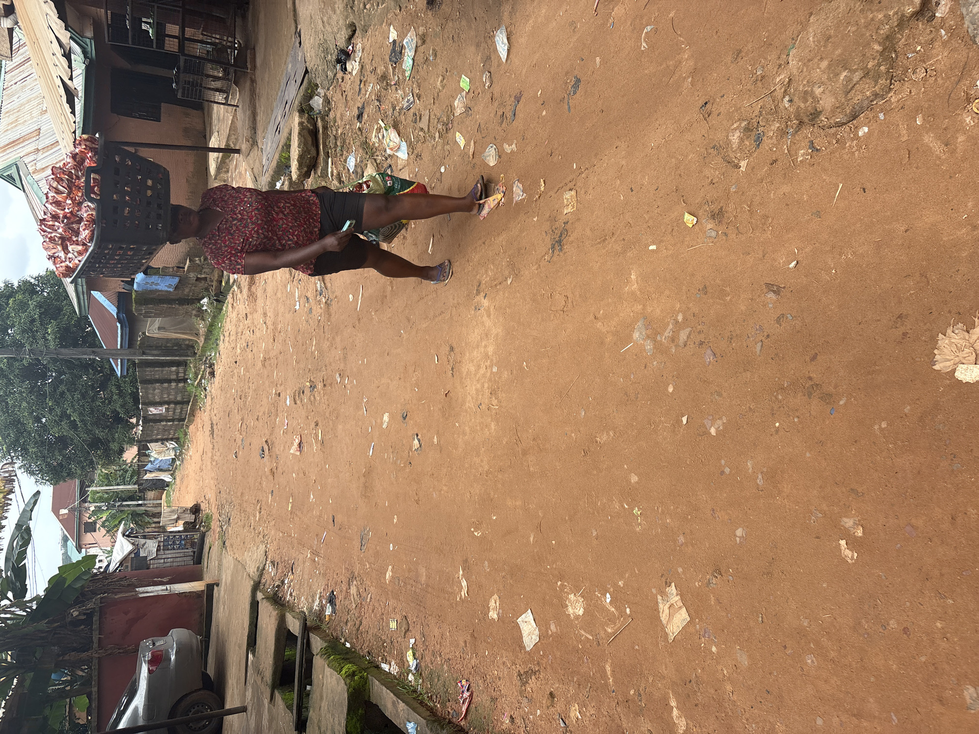 In Nkpor, Nigeria, a woman walks down a dusty, unpaved street, carrying a large black basket filled with red and white packaged goods on her head. She wears a red and white patterned short-sleeved shirt and dark shorts. Her left hand holds a green and white striped bag, and she wears dark flip-flops. The street is littered with debris and trash, including plastic bags, paper scraps, and discarded wrappers. To the left, a grey sedan is parked next to a red wall. Buildings with corrugated metal roofs line the right side of the street, and a stone wall runs along the middle. The sky is overcast.