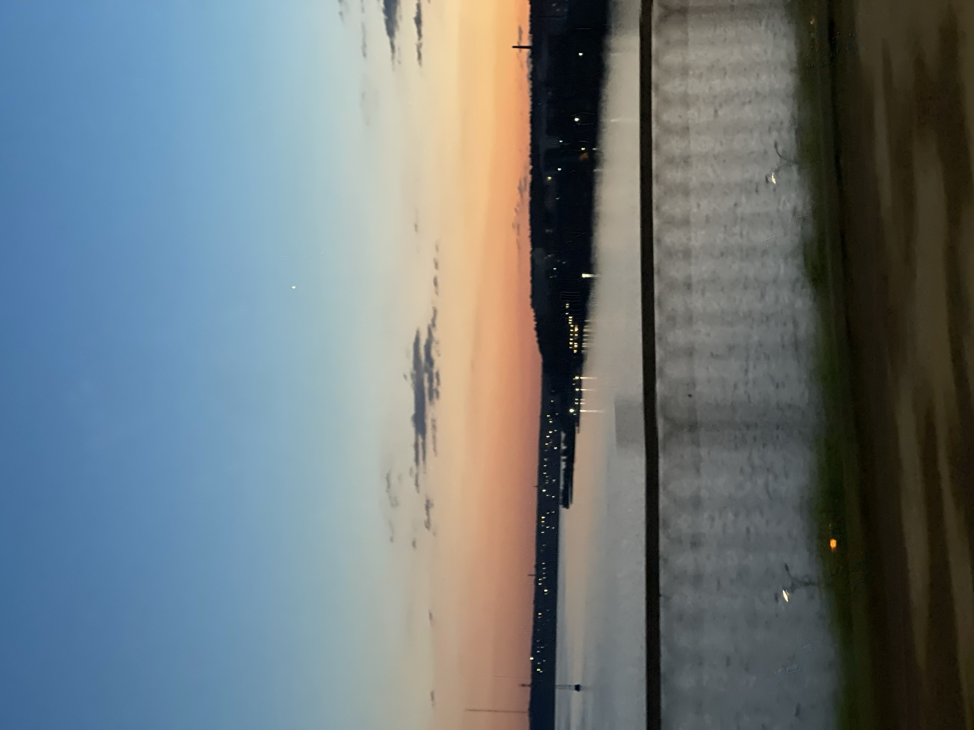The image shows a twilight scene over a body of water, likely a river or lake, with a city skyline in the distance. The sky transitions from a clear blue at the top to a warm orange and pink hue near the horizon, indicating the sun has recently set. Scattered clouds are visible in the sky. On the far bank, a dark silhouette of trees and buildings is punctuated by numerous small lights, suggesting a populated area. A thin, tall structure, possibly a radio tower or antenna, stands prominently on the left side of the image near the water's edge. The water itself appears calm, reflecting the sky and the distant lights. In the foreground, a blurred railing or barrier separates the viewer from the scene, with a dark, out-of-focus foreground suggesting a road or pathway. The overall atmosphere is serene and tranquil, capturing the transition from day to night.