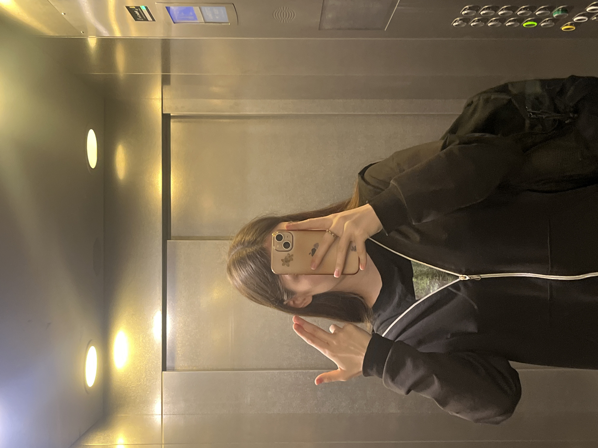 A young person with shoulder-length brown hair takes a selfie in an elevator. They are wearing a black zip-up hoodie over a black t-shirt, and have a black backpack slung over their right shoulder. Their left hand is raised, forming a "gun" shape with their fingers, and their right hand holds a mobile phone to capture the reflection. The elevator has metallic walls and ceiling with recessed circular lighting. On the right wall, an elevator control panel with illuminated buttons is visible. The context does not provide specific information about the location in Saint Petersburg, Russia.