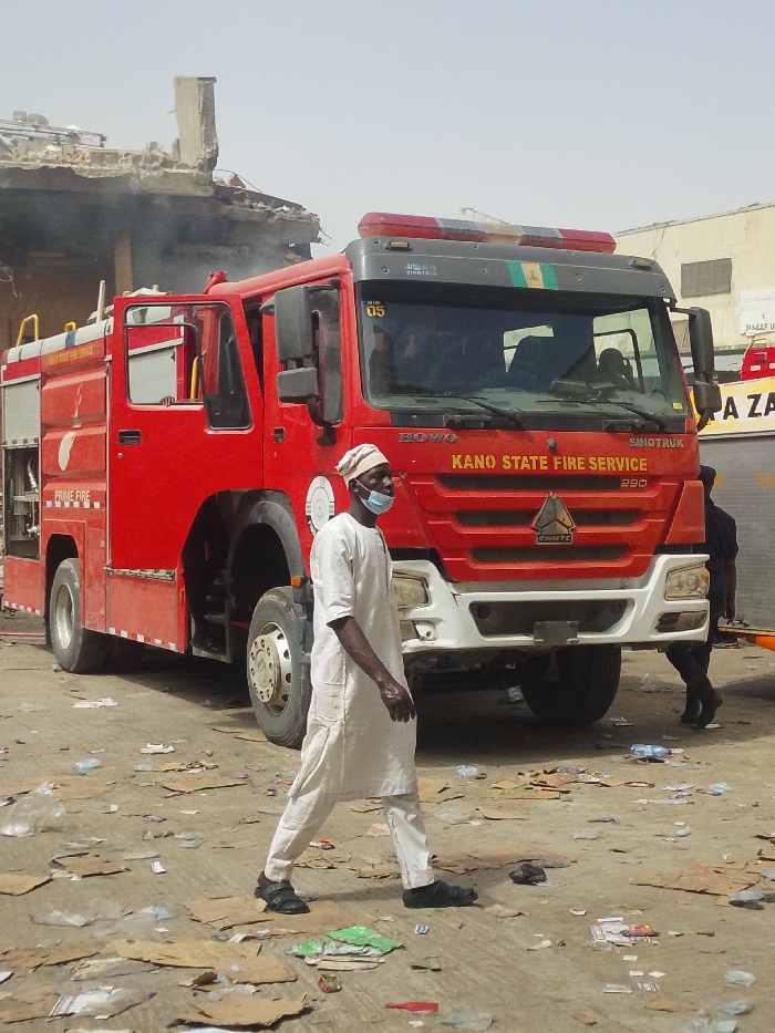 This image captures a daytime scene in Fagge C, Nigeria, depicting the aftermath or ongoing response to an incident. A large, red "KANO STATE FIRE SERVICE" Sinotruk HOWO 290 fire engine, marked with "PRIME FIRE" on its side, is prominently parked, facing slightly towards the viewer. Smoke is visible emanating from a severely damaged or partially collapsed concrete structure in the upper left background. In the foreground, a dark-skinned male subject walks from left to right. He wears a light-colored tunic and trousers, a light-colored cap, and a blue surgical mask pulled down under his chin, along with dark sandals. The ground is unpaved, extensively covered with various debris, including cardboard pieces, plastic bottles, and wrappers. Another individual in dark attire is partially visible behind the fire truck on the right side. The overall environment suggests an urban area affected by a fire or structural failure.