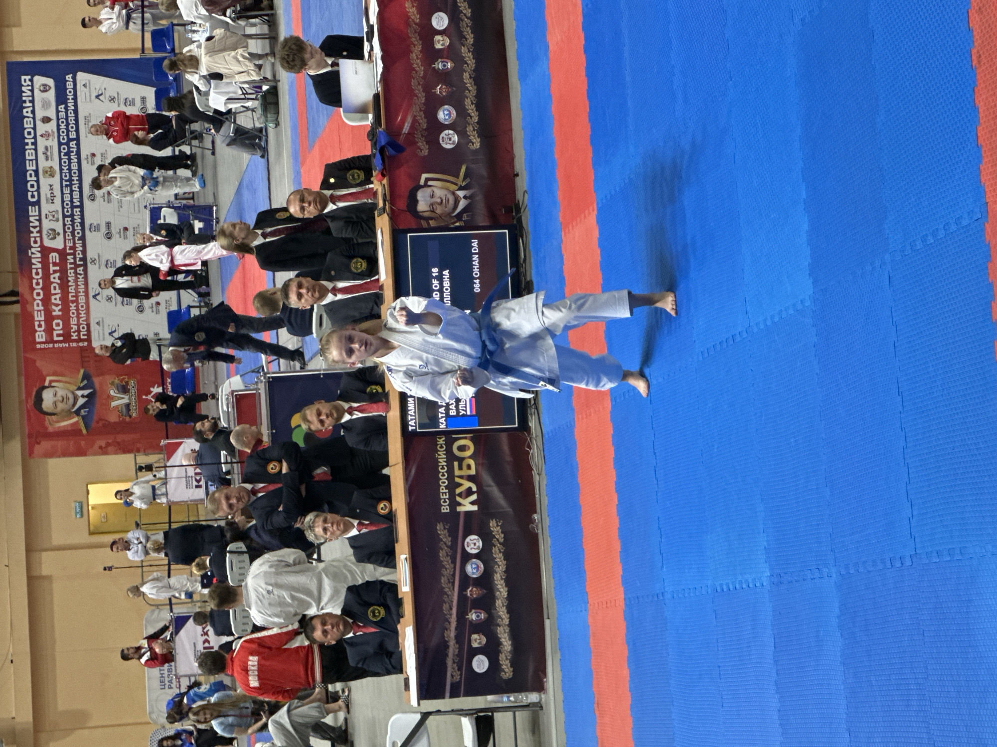 The image shows a karate competition in Demenshchina, Russia. A young woman wearing a white karate gi with a blue belt is performing a kata on a blue and red mat. A screen in the background reads "064 OHAN DAI," indicating the name of the kata. Spectators and judges are seated in the background. Banners displaying the event's name, "Всероссийские Соревнования по Каратэ" (All-Russian Karate Competition), and "Кубок Памяти Героя Советского Союза Полковника Григория Ивановича Бояринова" (Cup in Memory of Hero of the Soviet Union Colonel Grigory Ivanovich Boyarinov), are also visible.
