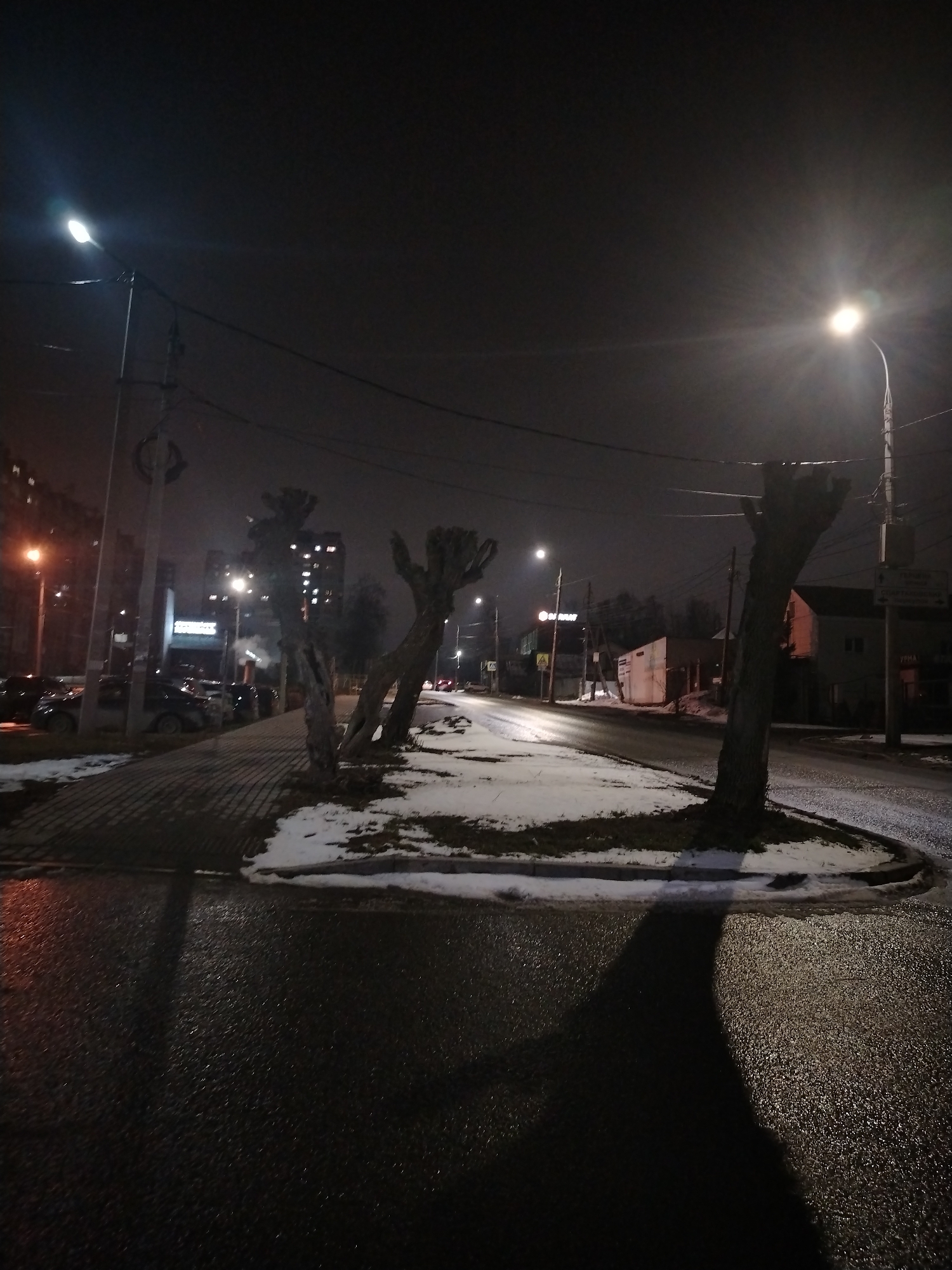 This is a nighttime view of a street in Izhevsk, Russia. The scene is illuminated by streetlights, their light reflecting on the wet road surface. There is snow remaining on the ground, particularly on the raised median along the street. Several bare, heavily trimmed trees stand on this median. Cars are parked along the left side of the street, and buildings are visible in the background, including apartment buildings and smaller structures. Power lines are overhead. The overall impression is one of a cold, wet winter night in an urban area.