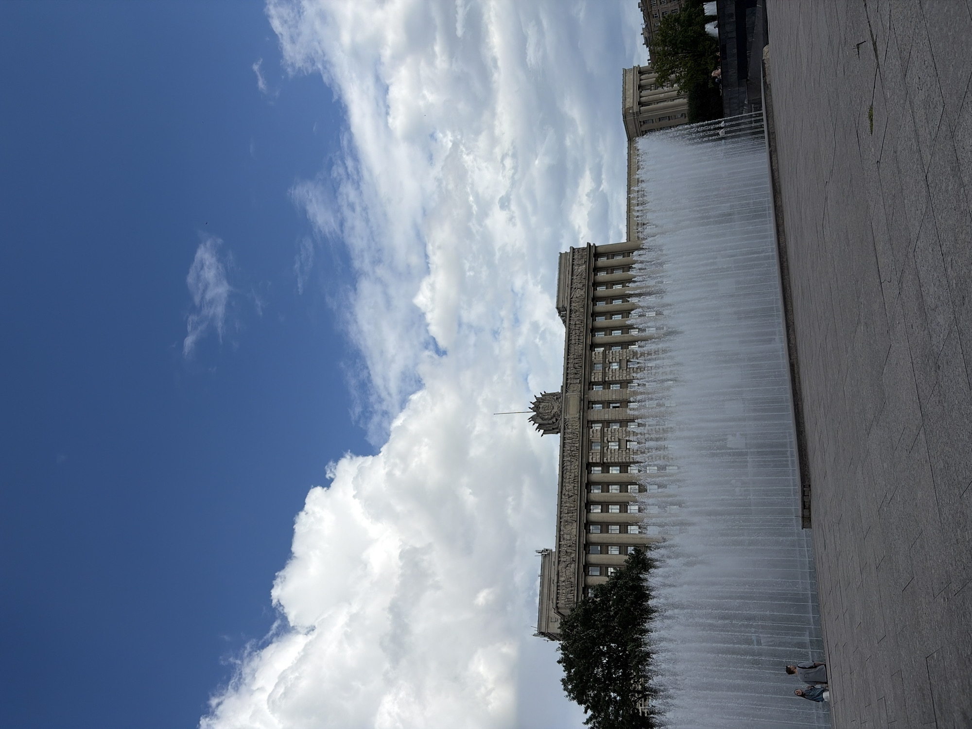 This is a photograph of the Moscow Triumphal Gate in Saint Petersburg, Russia, viewed from across a large fountain. The gate is a grand, neoclassical structure with a long facade, featuring rows of windows, columns, and ornate sculptures along its upper level. A large fountain is in the foreground, with water spraying upwards in a dense sheet, obscuring much of the lower portion of the gate.
In front of the fountain, on a stone plaza, two people are standing and facing away from the camera. They are relatively small in the frame, with one person wearing a light-colored jacket and the other a darker one. The plaza is paved with large, grey stone slabs.
The sky above is a vibrant blue, dotted with large, fluffy white clouds, particularly on the left side of the image. The overall lighting suggests a bright, possibly late afternoon or early morning day. The scene is calm and expansive, with the impressive architecture of the gate dominating the background.