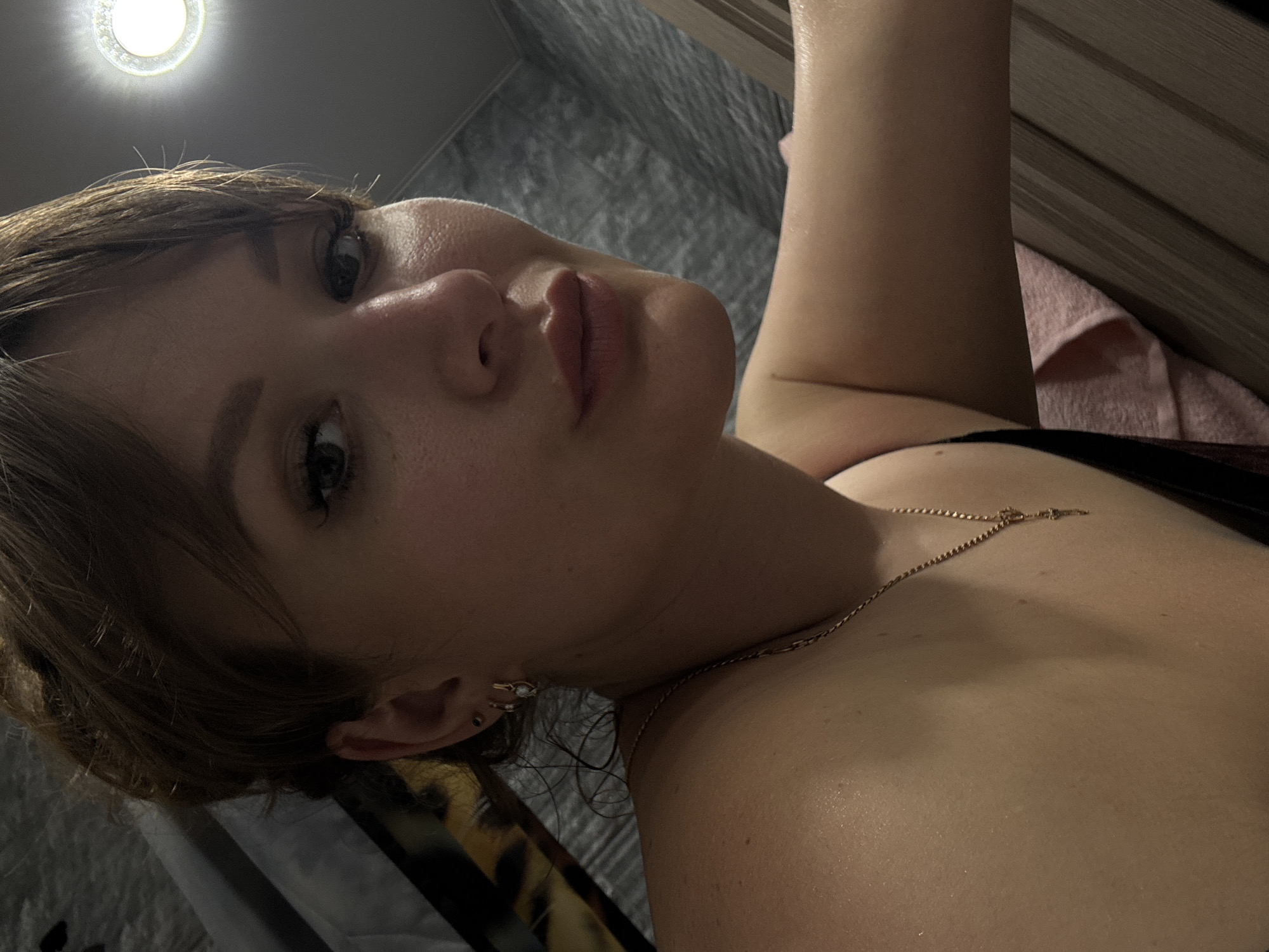 A close-up, angled shot of a woman with light skin. She has brown hair and is wearing a gold necklace. Her face is tilted, and she is looking at the camera. In the background is a grey wall and a light source. The location is indoors, but not identifiable as Chaikovsky, Russia.